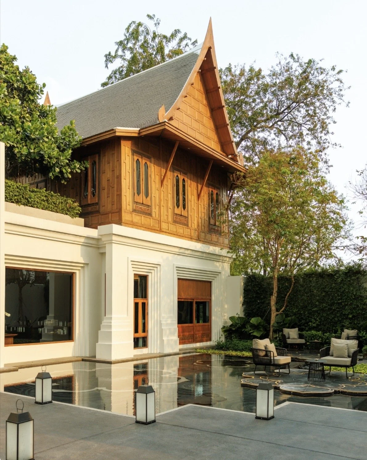A two-story house with a traditional Thai-style wooden upper level and a modern white lower level. The house features a high, steeply pitched roof with ornate gable details. In front, there is a patio with outdoor seating, a small pool or water feature, and decorative lanterns. The landscape includes trees and lush greenery surrounding the house.