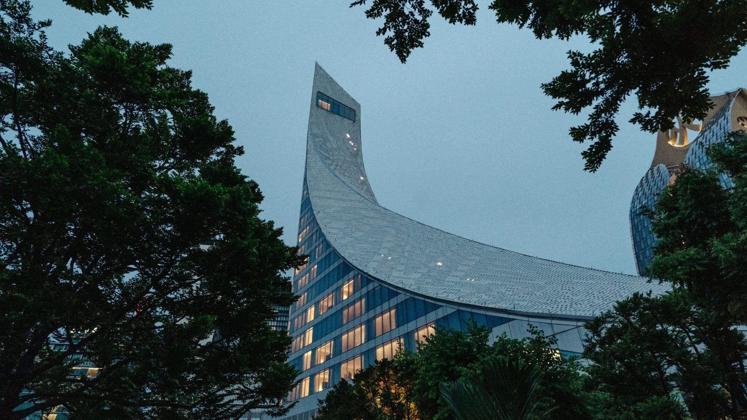 Tall modern skyscraper surrounded by green trees, with windows illuminated at dusk.