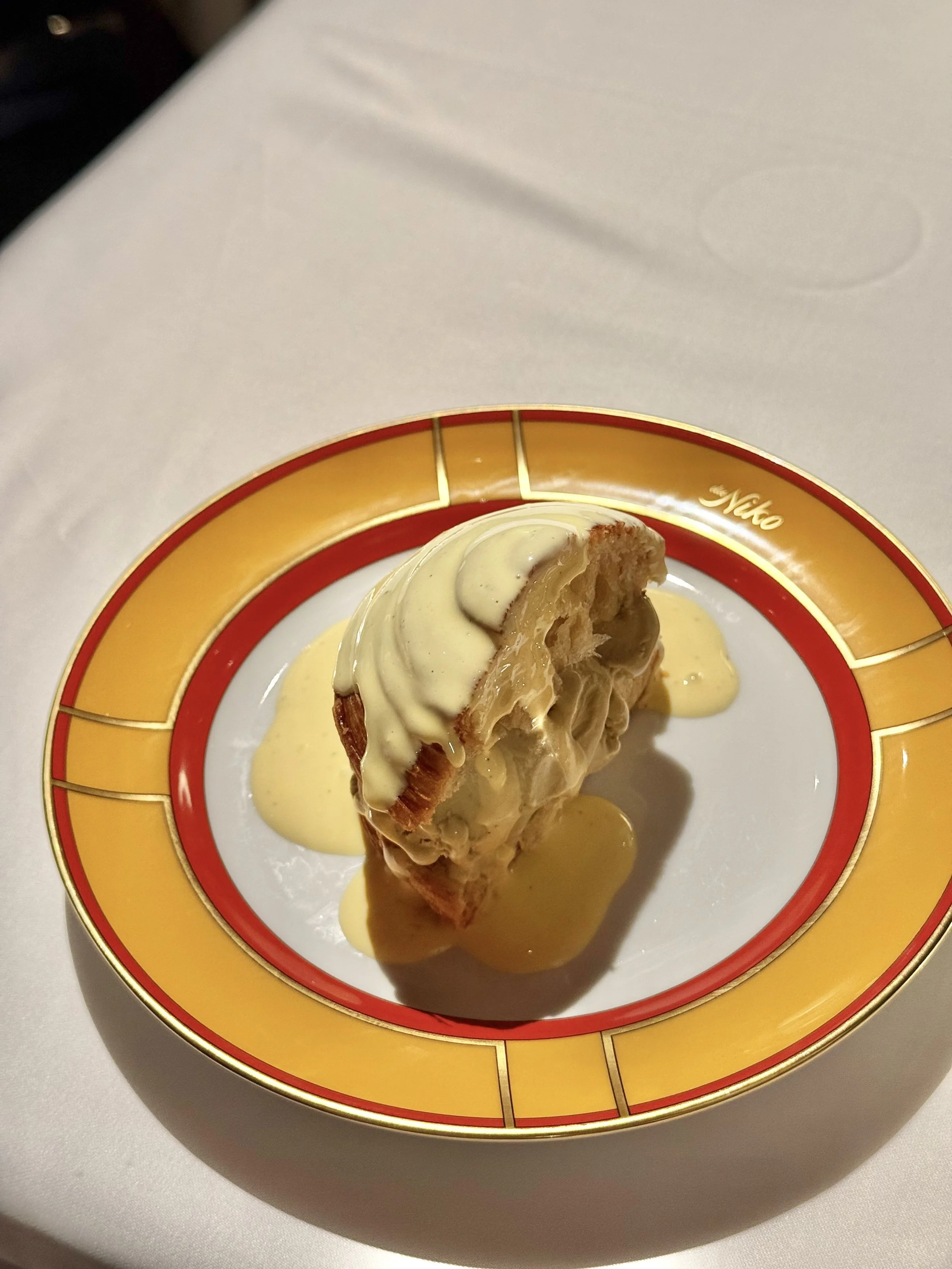 Half a flaky brioche filled with pistachio gelato and topped with crema inglese on a yellow, red and gold plate.