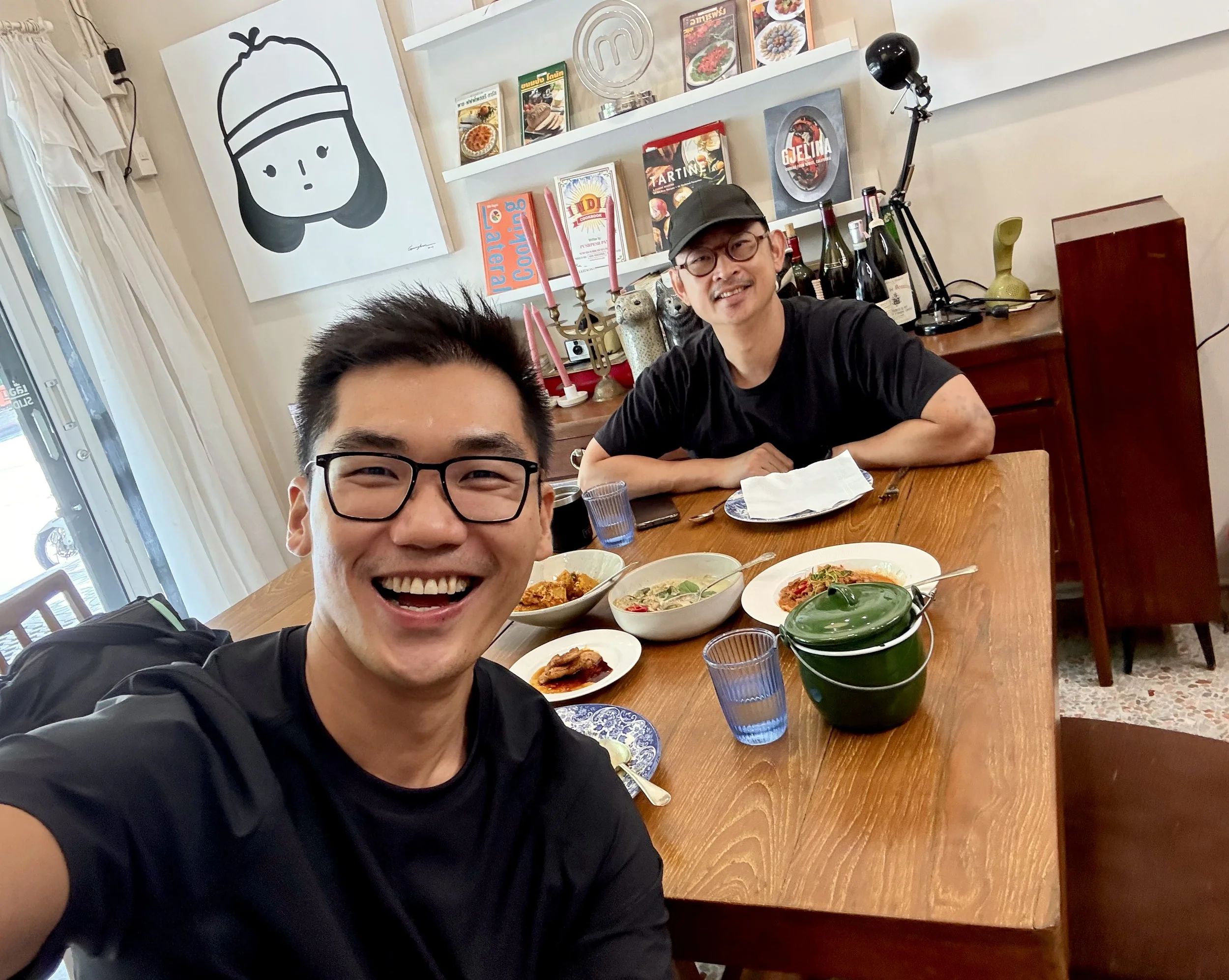 JJ Wong and Angkrit smiling across a wooden table set with three curries, a plate of pork chop and blue drinking glasses.