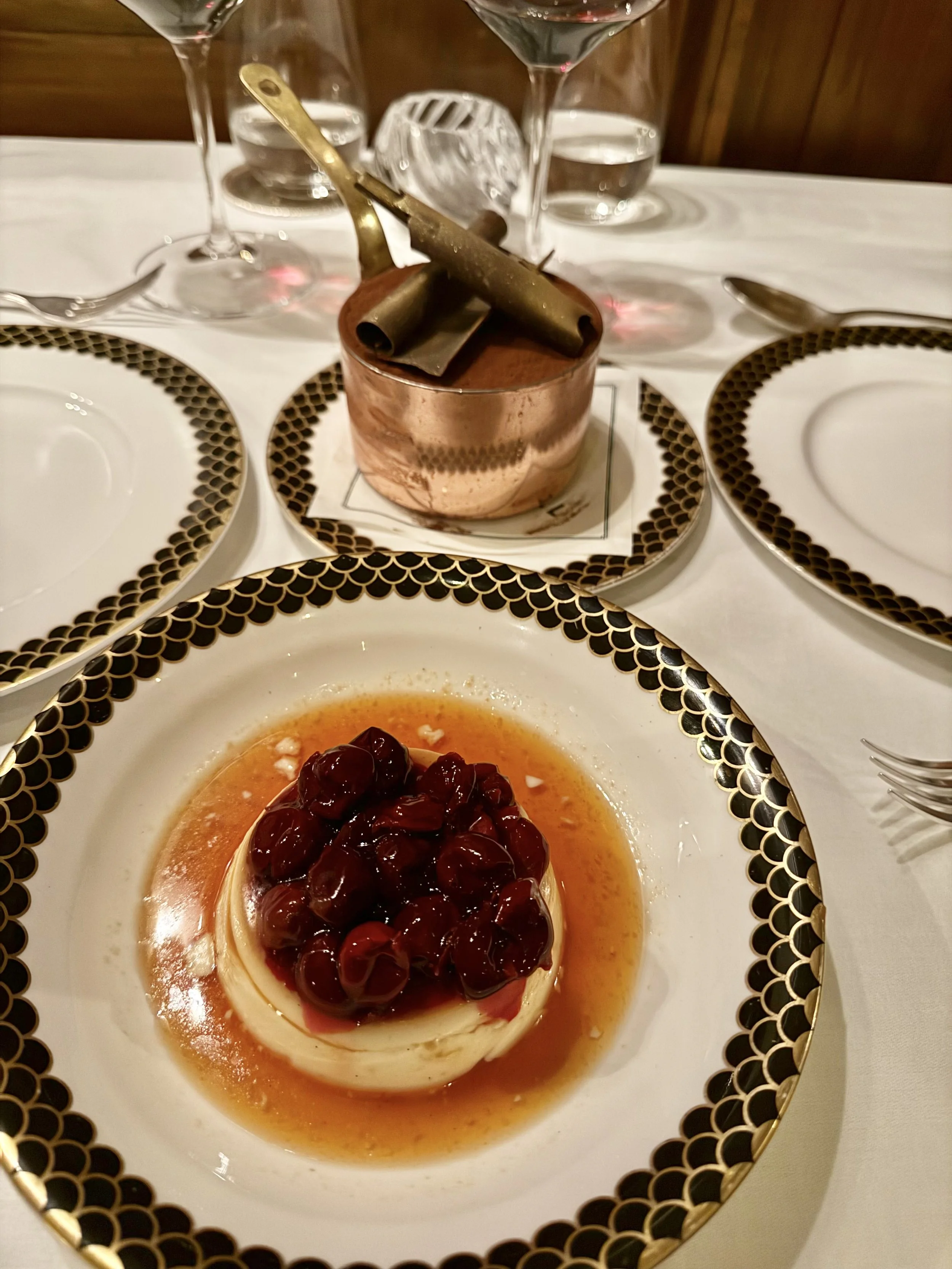 Crème caramel topped with cherries in the foreground, with chocolate mousse topped with chocolate curls in a copper pot behind it on the white tablecloth at Lutyens Grill.