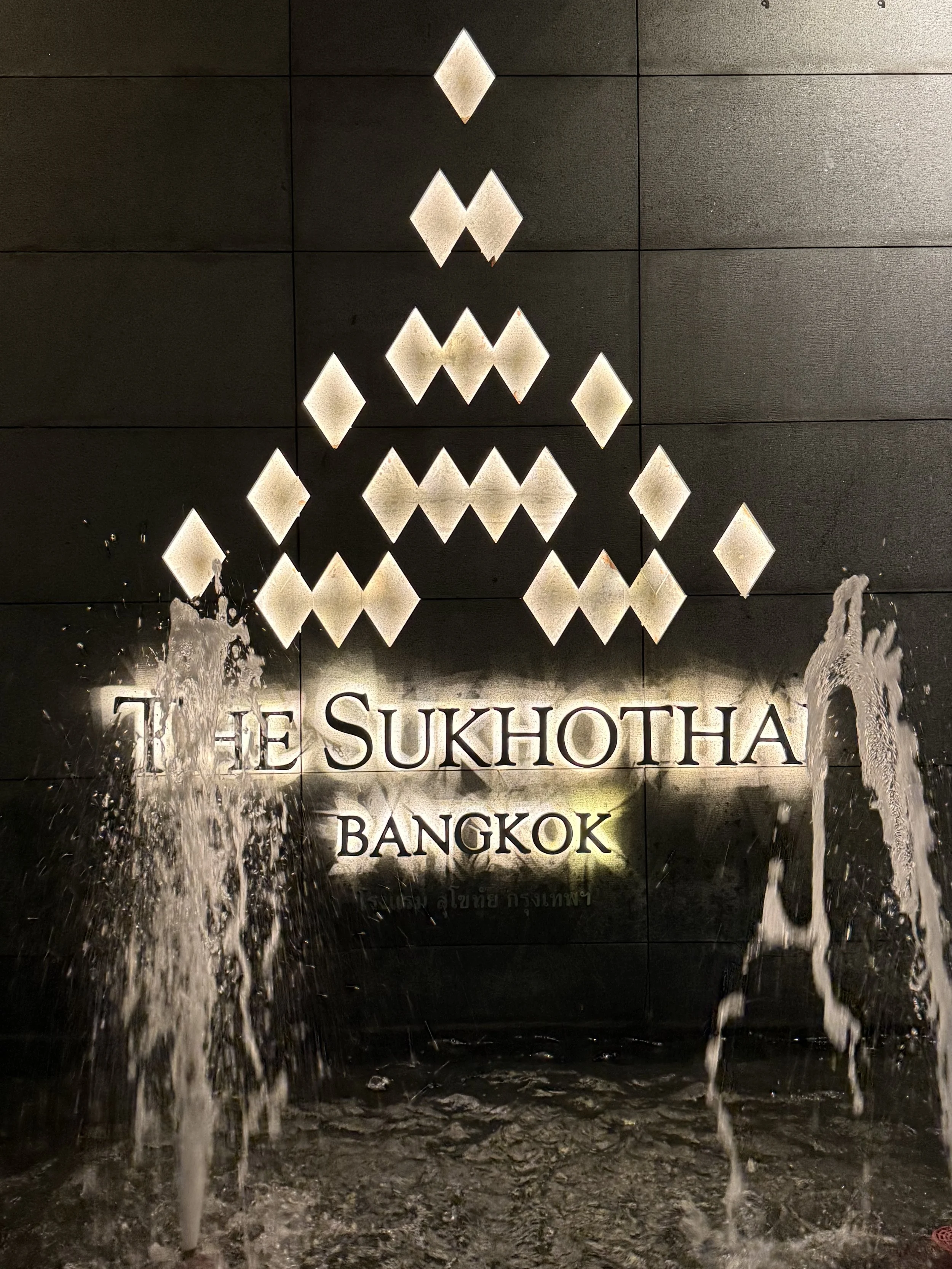 Illuminated diamond shapes form The Sukhothai Bangkok emblem above the hotel name and a water feature.
