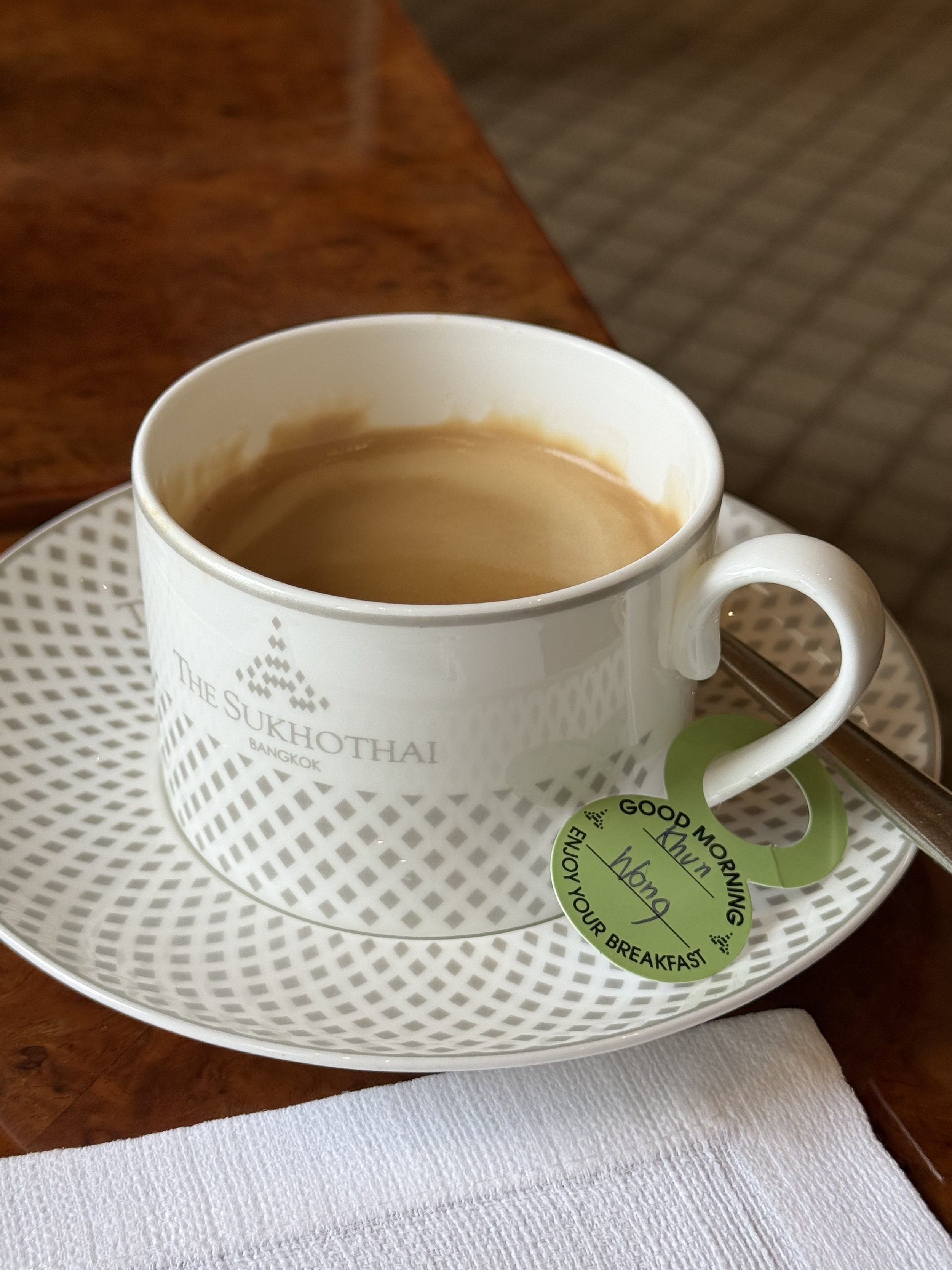 A cup of coffee on patterned porcelain at The Sukhothai Bangkok, with a small green greeting note on the saucer.