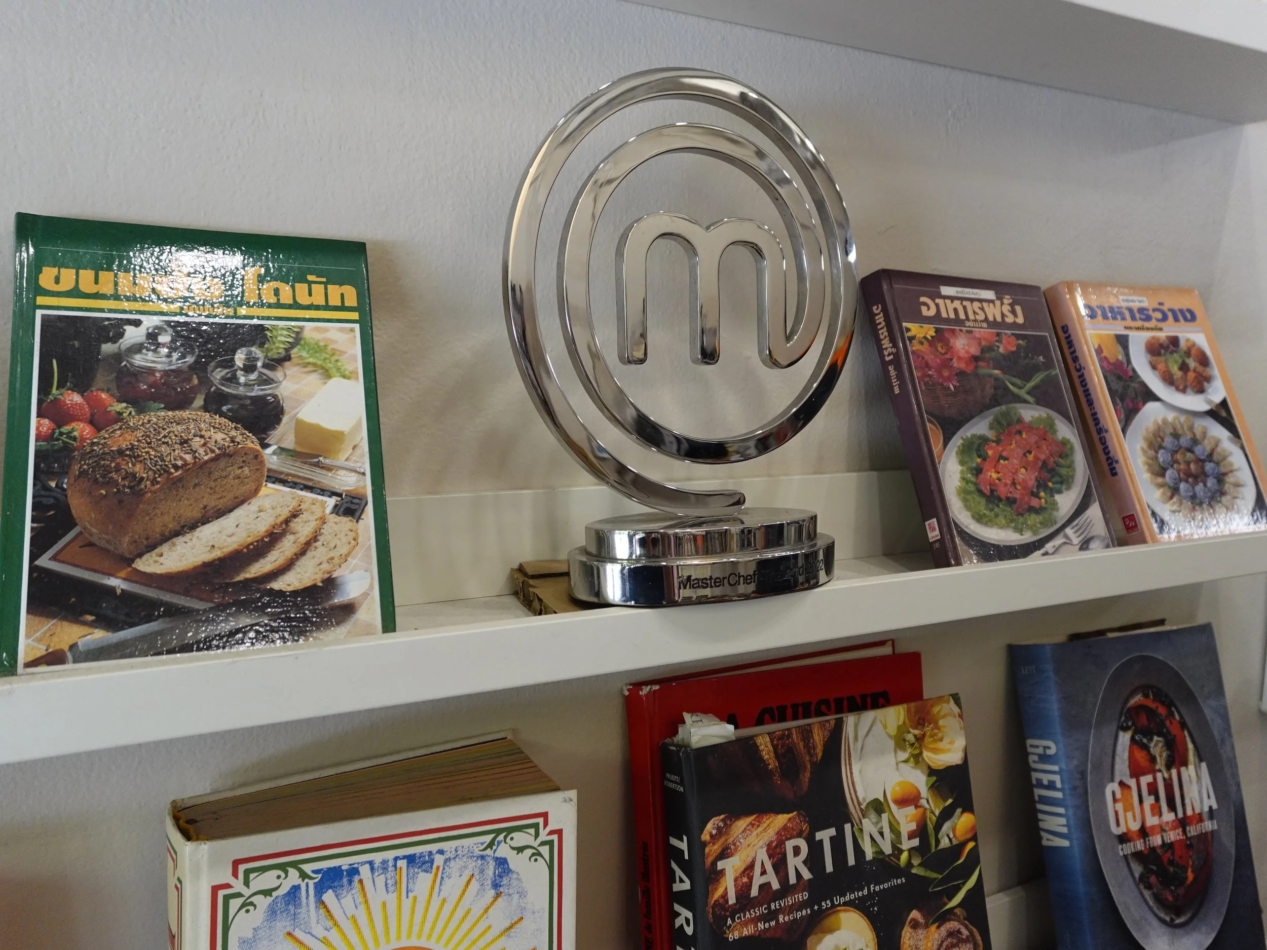 Silver MasterChef trophy on a white shelf between Thai cookbooks, with Tartine and Gjelina on the shelf below.