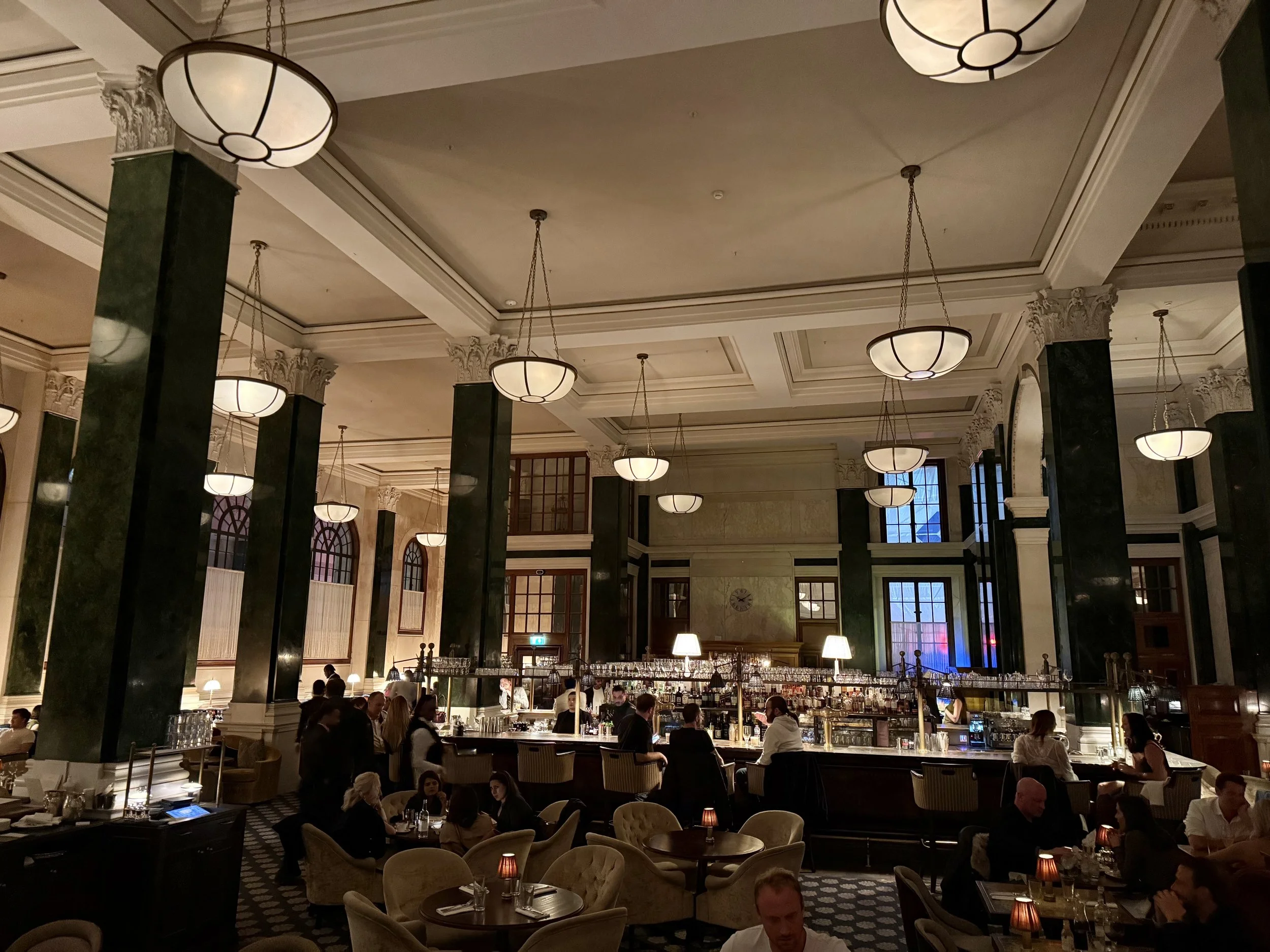 Evening view of Millie’s Lounge at The Ned, with people seated around lamp-lit tables and a long bar beneath tall green columns and bowl-shaped pendant lights.