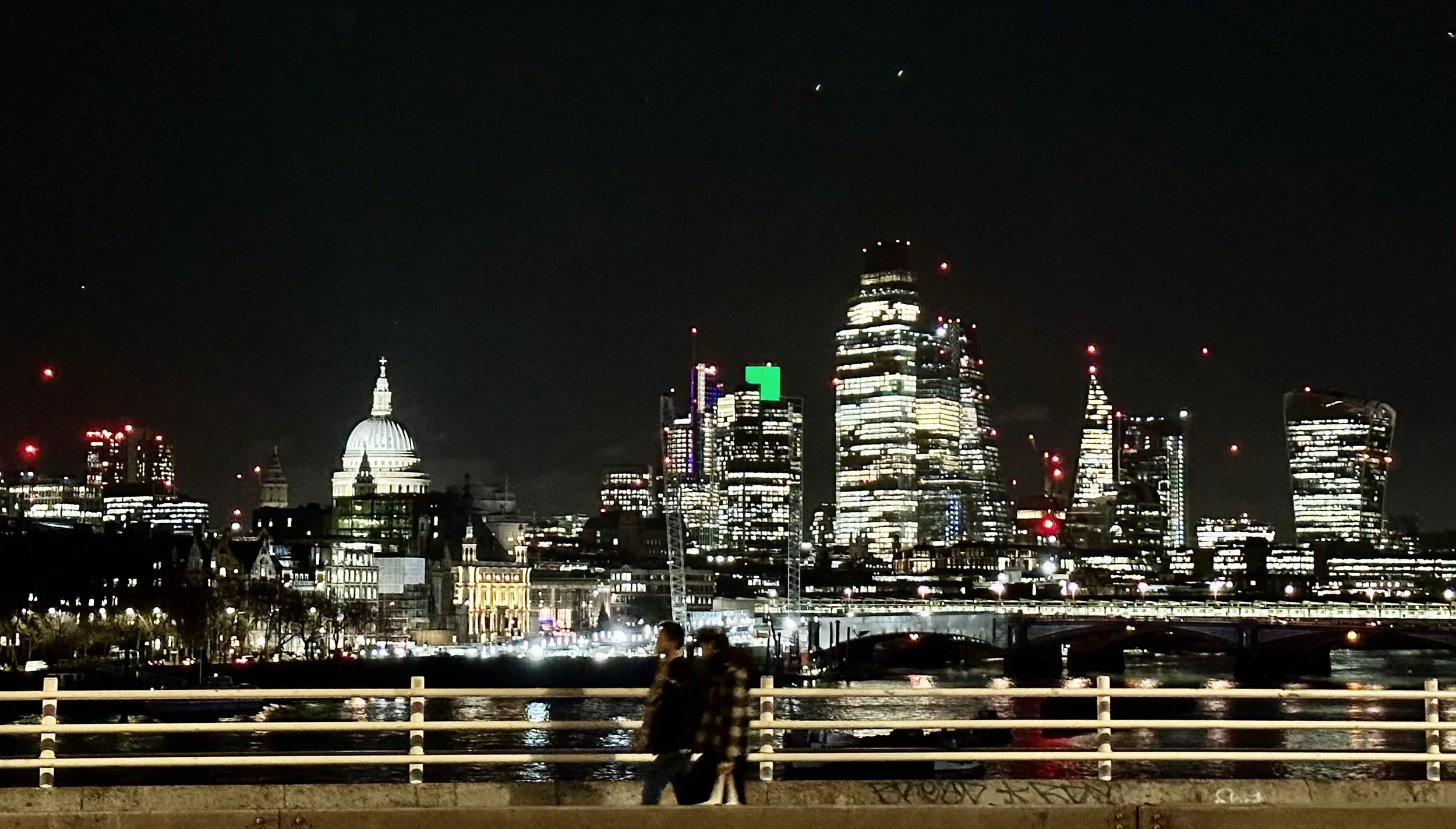 Nighttime city skyline with illuminated tall skyscrapers and a historic building with a dome, viewed across a river, with two people walking in the foreground.