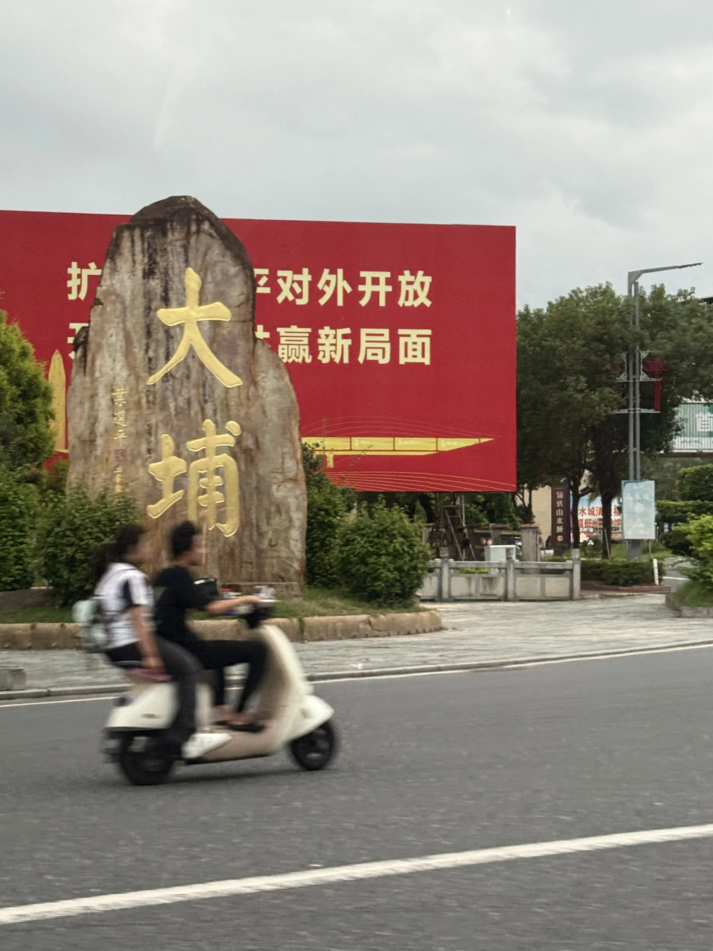 A roadside stone marker bearing the large gold Chinese characters 大埔, Dabu, as a scooter passes in front.