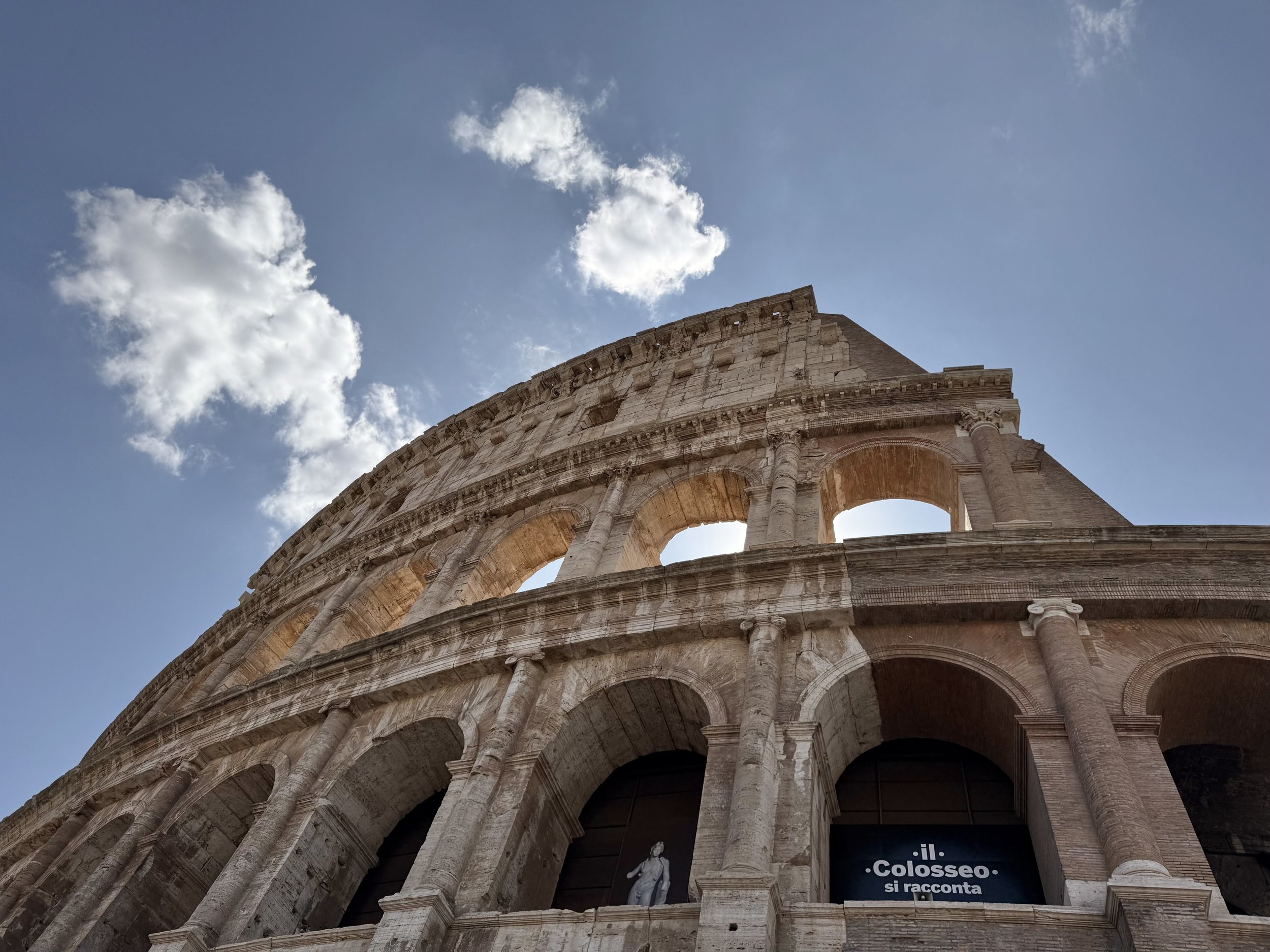 The image shows an upward view of the Colosseum's exterior with stone arches and columns, against a bright blue sky with scattered white clouds.