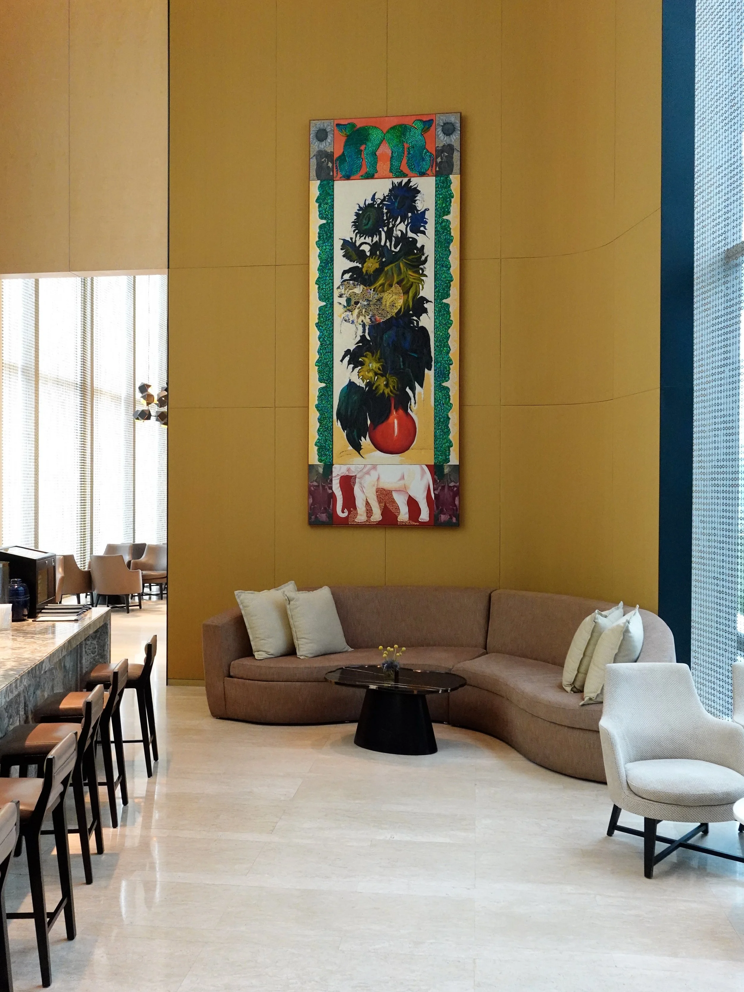 A curved beige sofa beneath a tall floral artwork, beside a stone bar counter and stools.