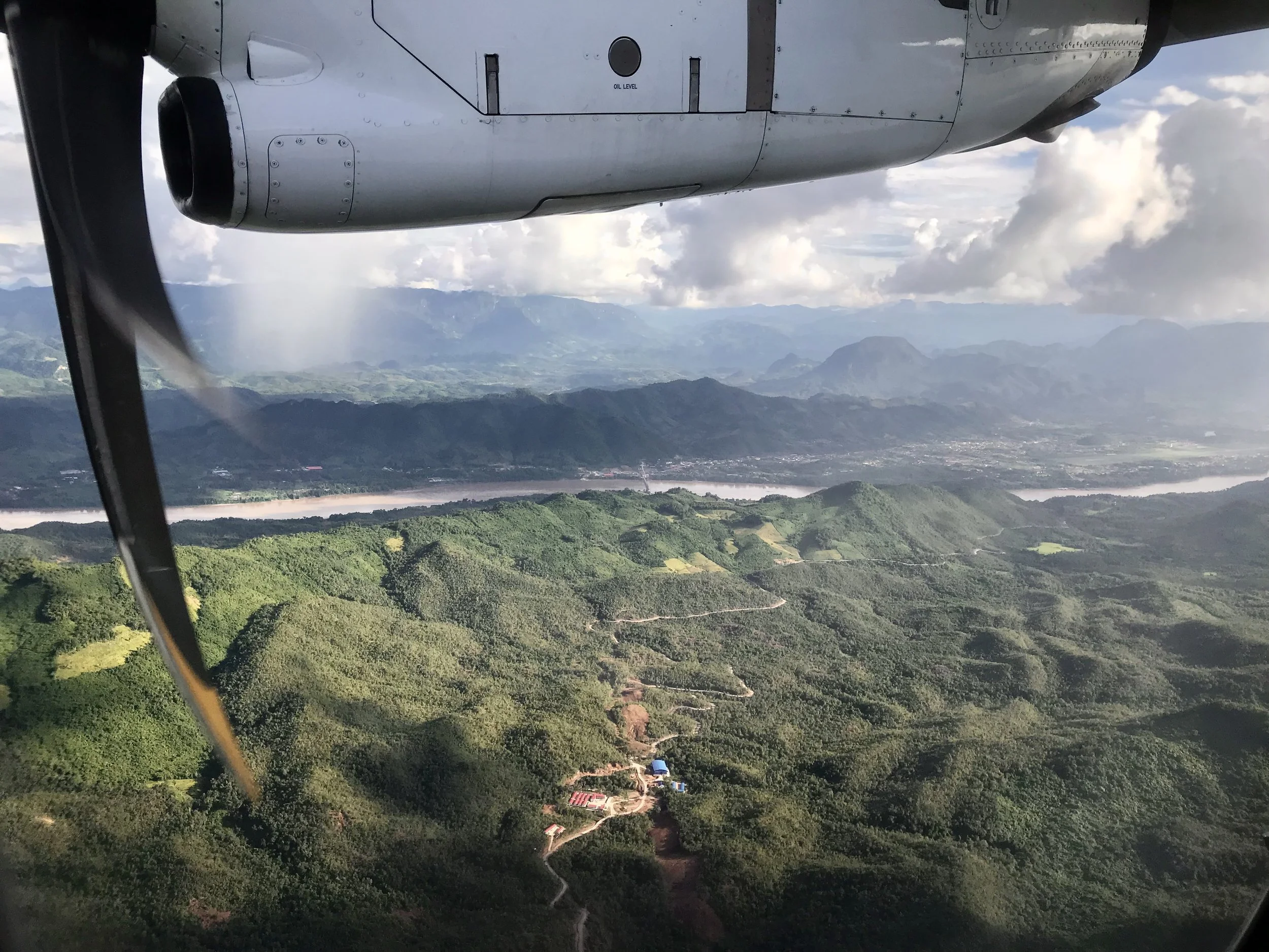 photograph taken through the window of a propeller aircraft: the silver engine and blurred propeller occupy the upper part of the frame, with green mountains, the Mekong and Luang Prabang below.
