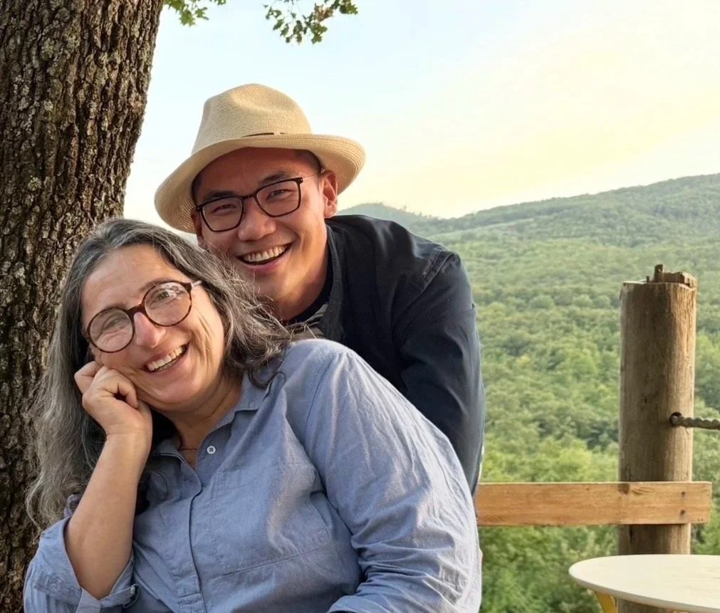Claudia Rizza and JJ Wong smiling together beside a tree on the wooden terrace above the vineyard.