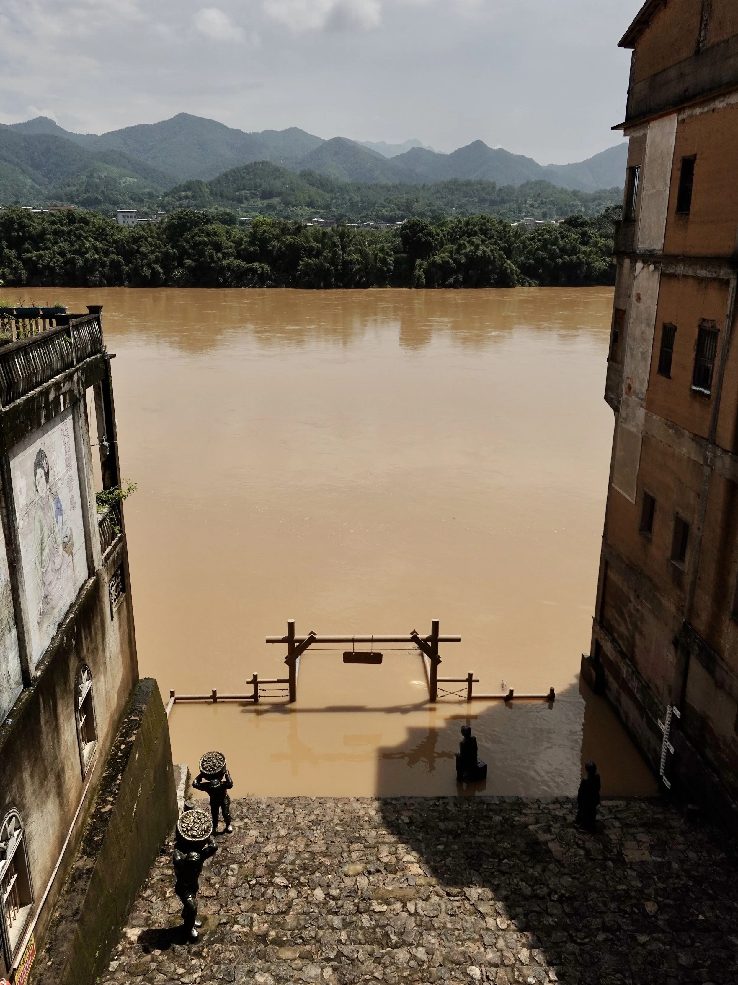 An elevated view of Huochuan Pier in Songkou, with muddy river water covering the lower landing, departure sculptures below and wooded hills beyond.