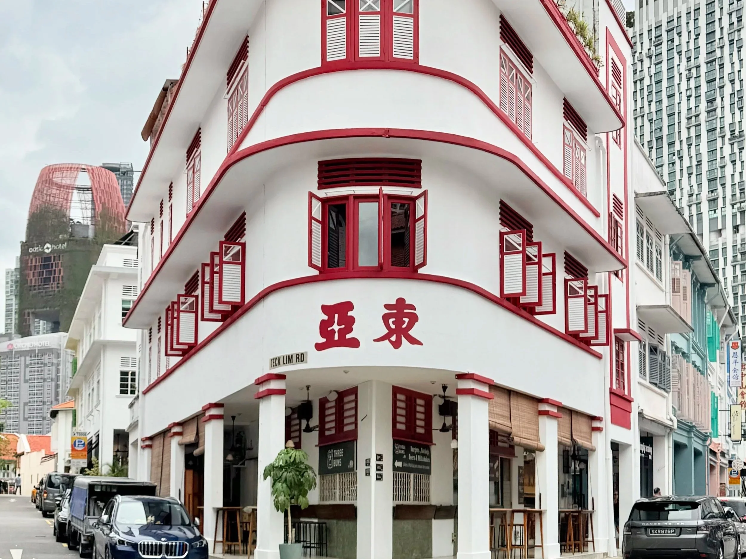 A white building with red accents and open window shutters, situated on a busy street with parked cars and high-rise buildings in the background.