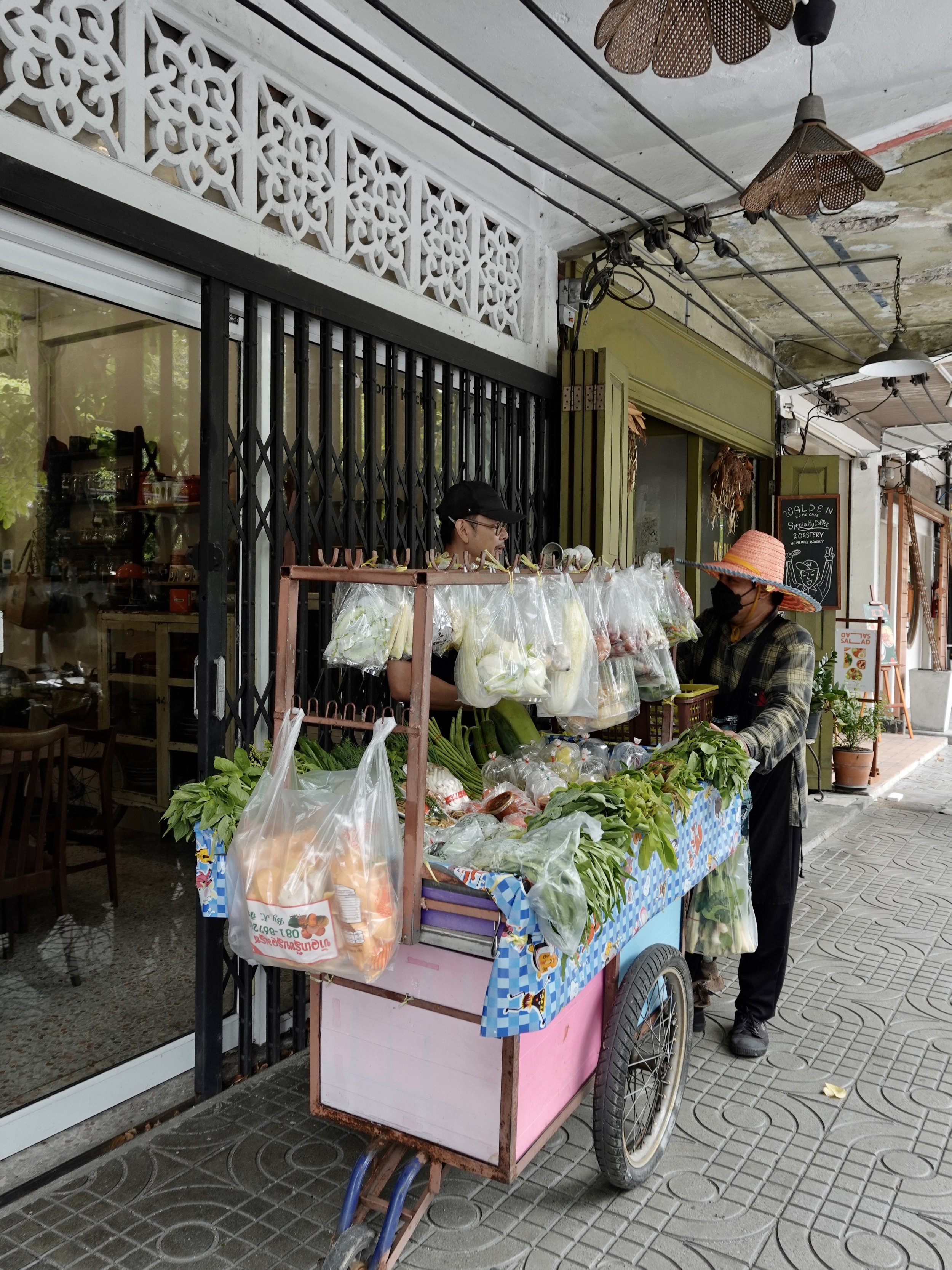 Angkrit and a vegetable vendor beside a pink wheeled cart outside a glass-fronted kitchen on a covered Bangkok pavement.