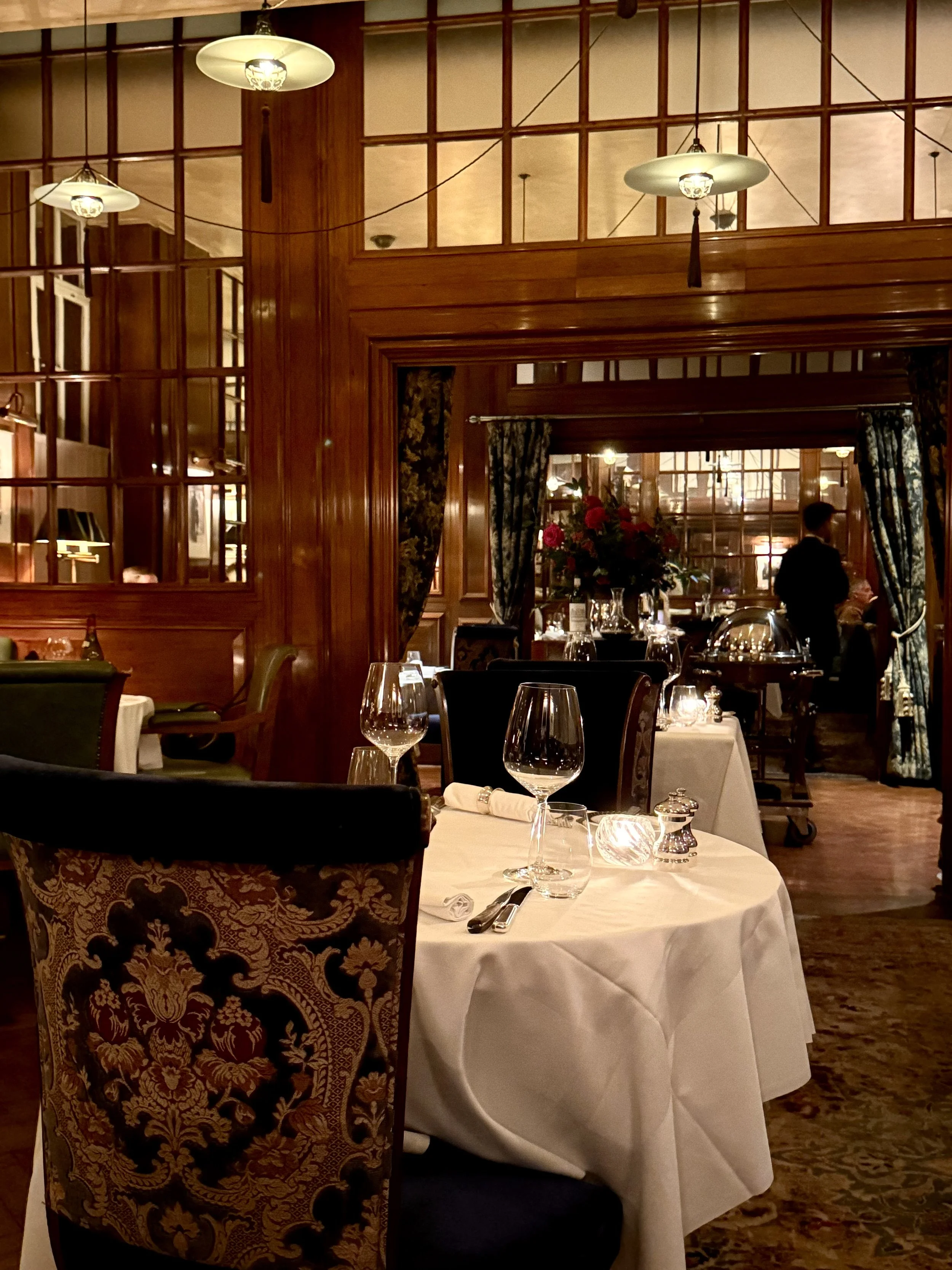 Lutyens Grill dining room at The Ned, with polished timber panelling, glazed partitions, pendant lights, patterned upholstered chairs and tables set with white linen and wine glasses.