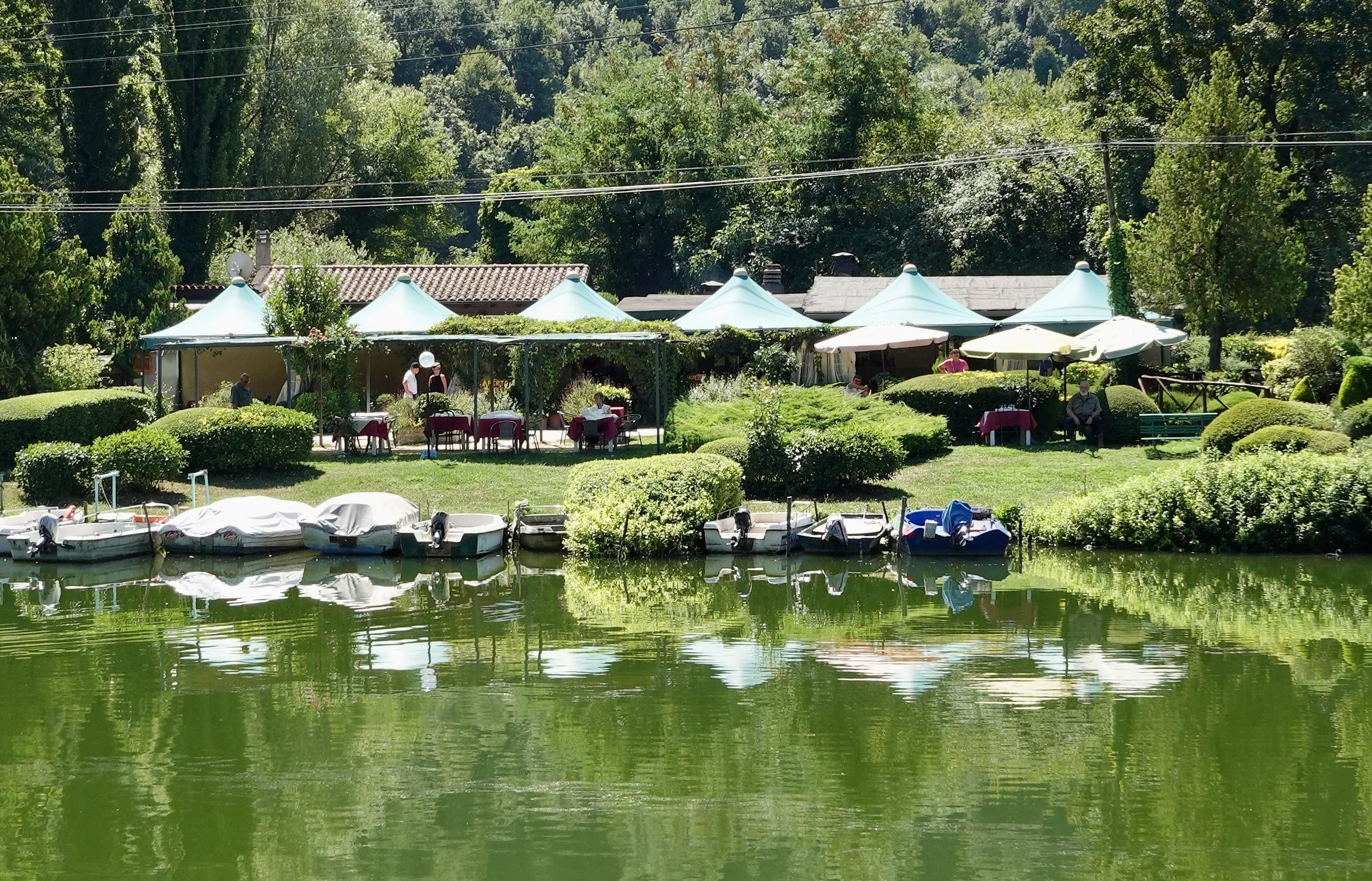 Green lake water reflecting a line of small moored boats, clipped shrubs, outdoor tables and pale turquoise canopies on the opposite bank.