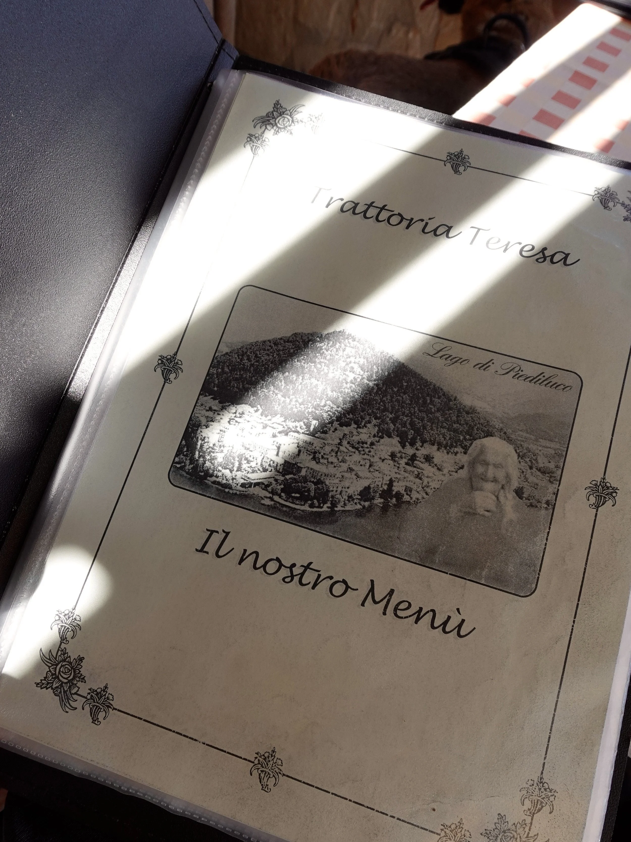 Angled close\-up of the menu in bands of sunlight and shadow\. The cover reads “Trattoria Teresa”, “Lago di Piediluco” and “Il nostro Menù”, with an old black\-and\-white image of the lake and a woman.