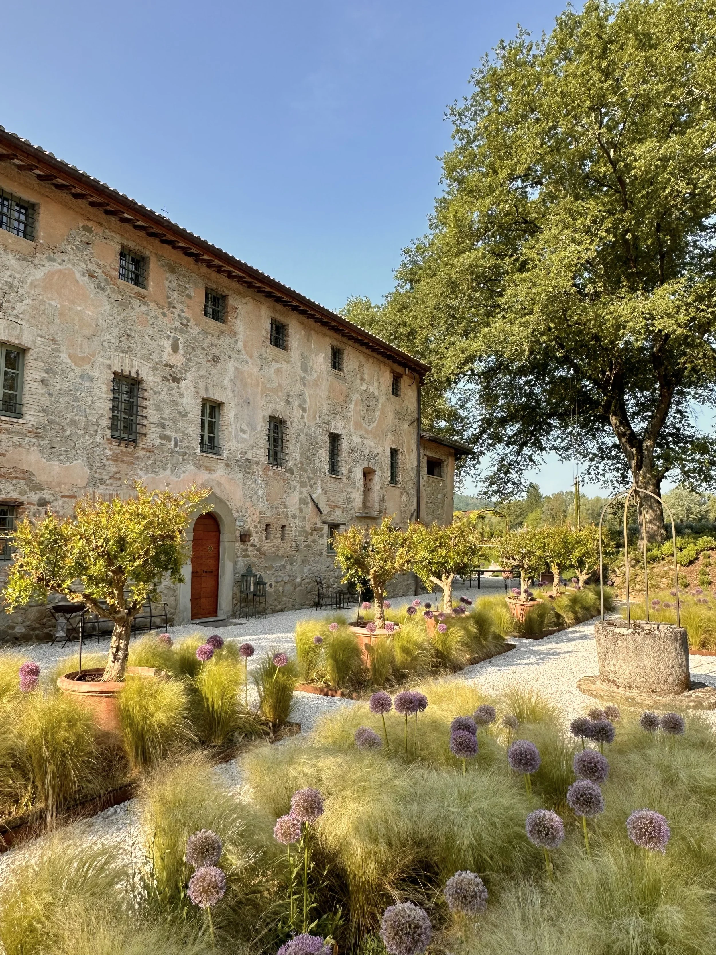 A rustic stone building with small barred windows, set adjacent to a well-maintained garden with potted trees, flowering plants, and decorative grasses, under a blue sky with a large tree on the right side.