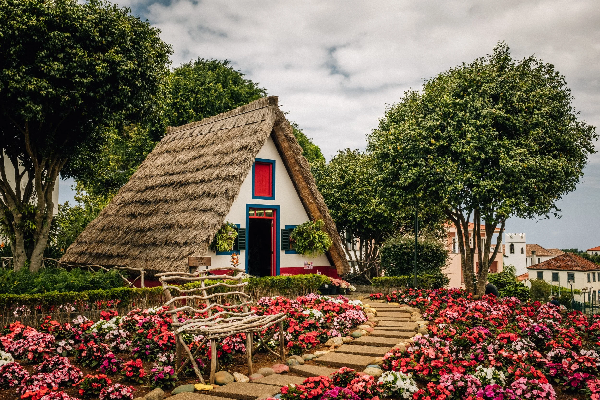 Colorful house with a thatched roof, red window and door, surrounded by vibrant pink, white, and red flowers and lush green trees.