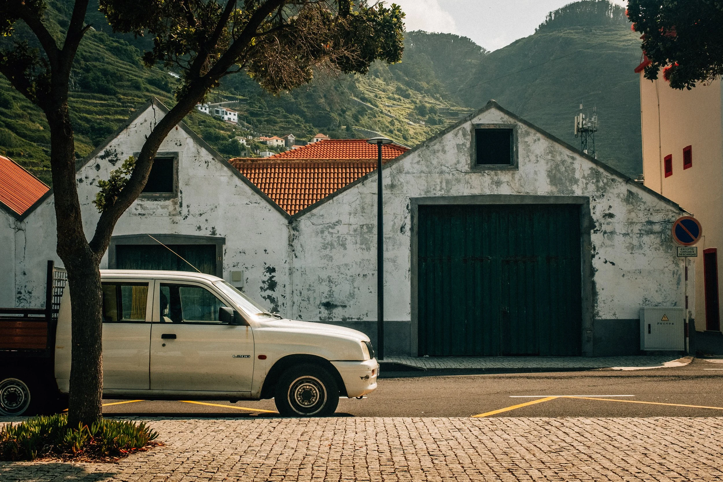 A white car parked on a paved street in front of an old, weathered building with a green garage door. Trees and hilly terrain with houses are visible in the background.