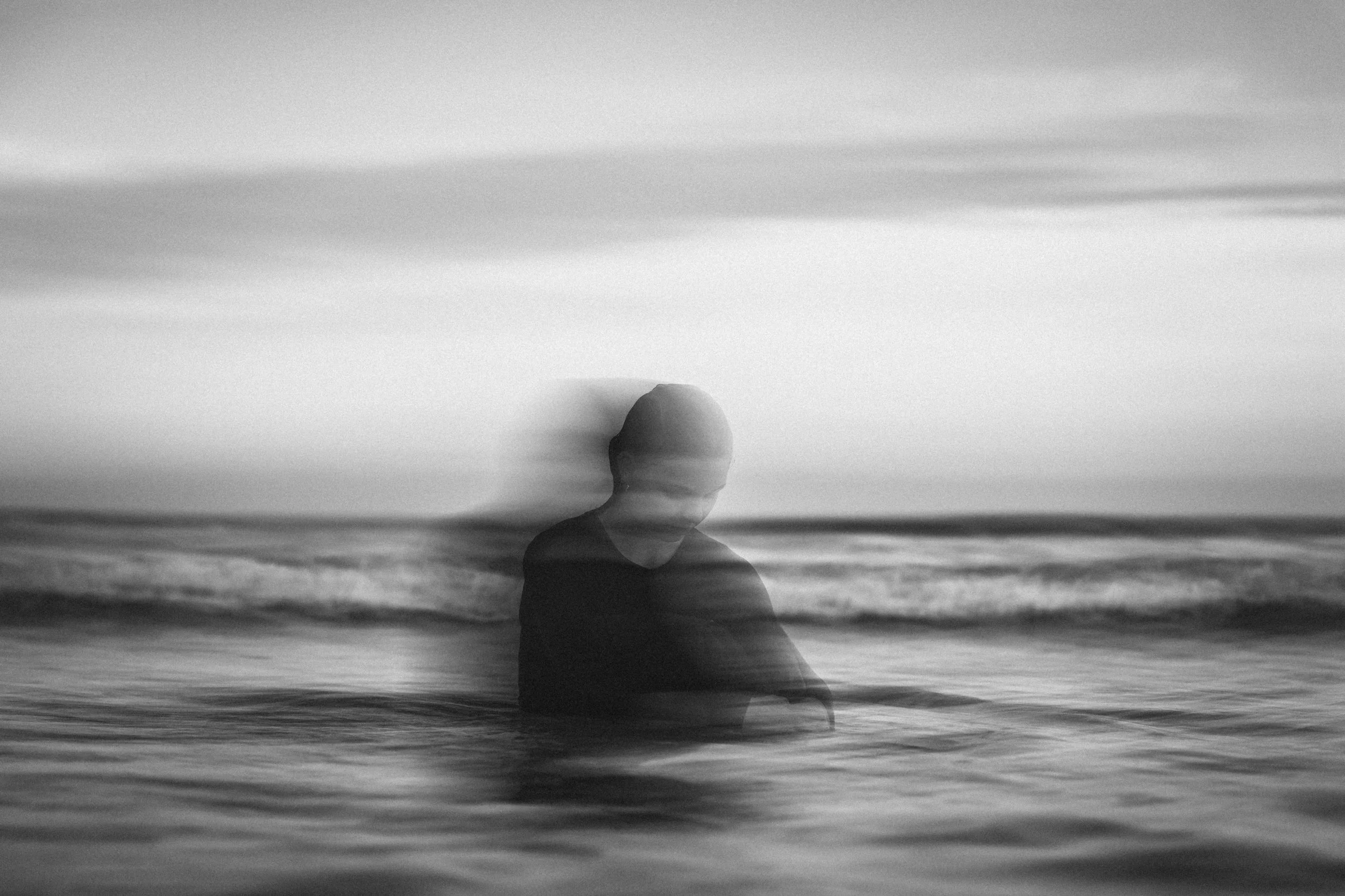A person is standing in the ocean at sunset or sunrise, with their face blurred or obscured, creating a dreamy, ethereal effect in black and white.