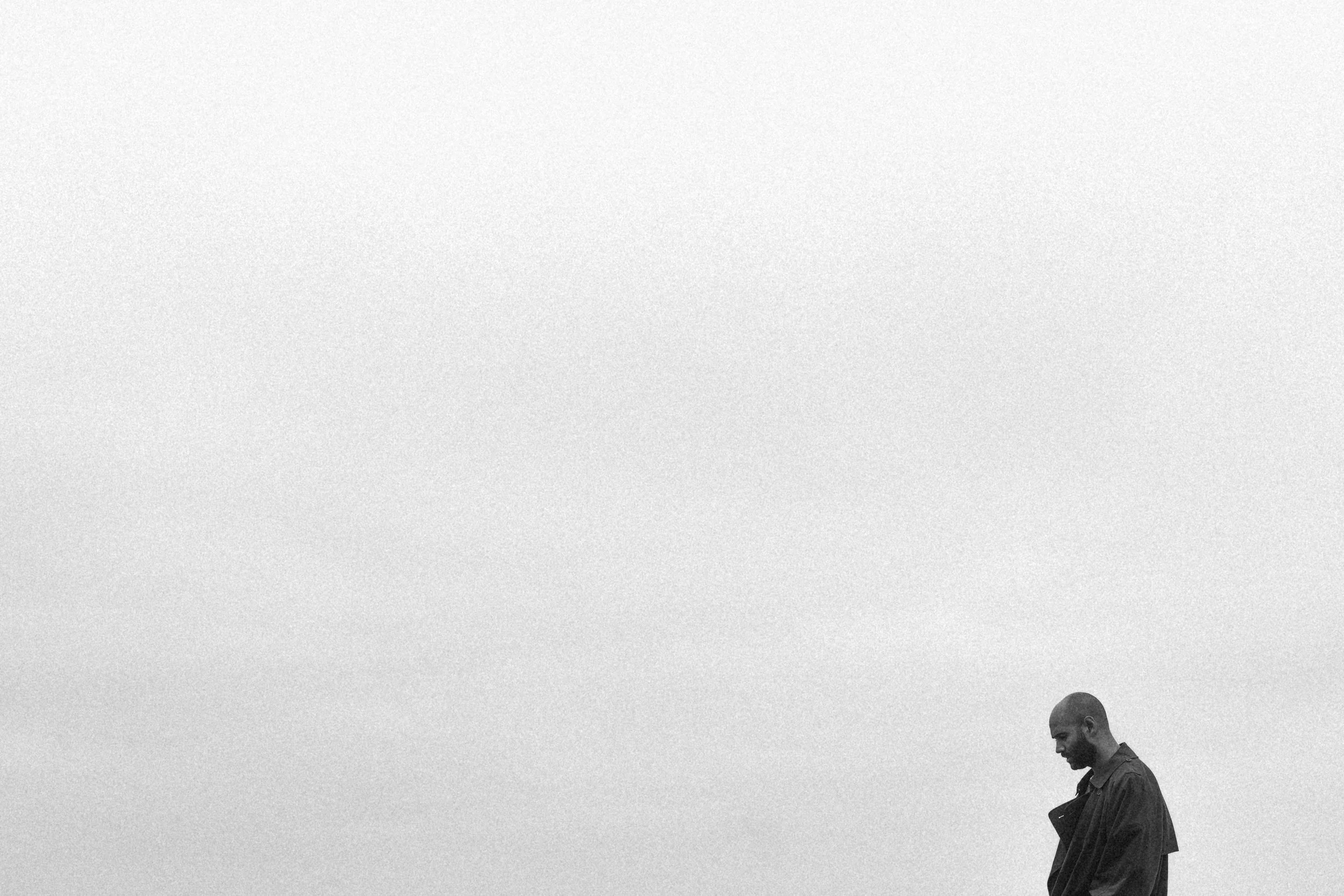 A black and white photo of a man with a beard looking down, standing outdoors against a cloudy sky background.