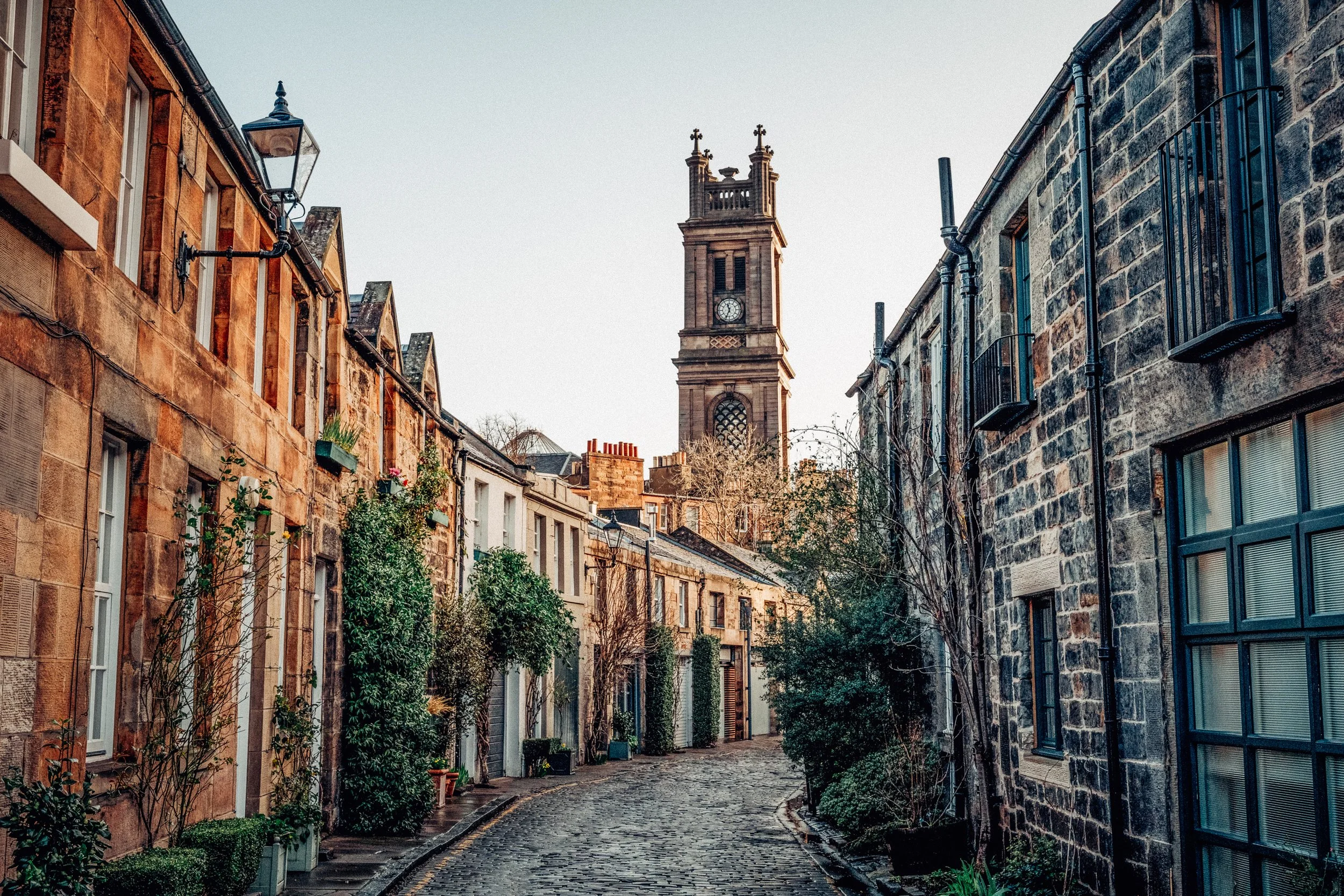 A narrow cobblestone street lined with old brick and stone buildings, with a clock tower in the background under a clear sky.
