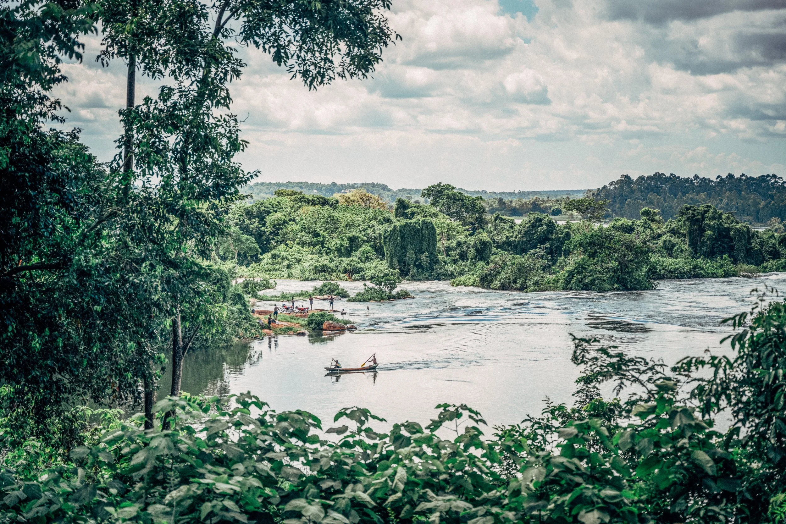 A river flowing through a lush green forest with trees on both sides and a partly cloudy sky above. Two people are in a small boat on the river, and area in the foreground is framed by branches and leaves.