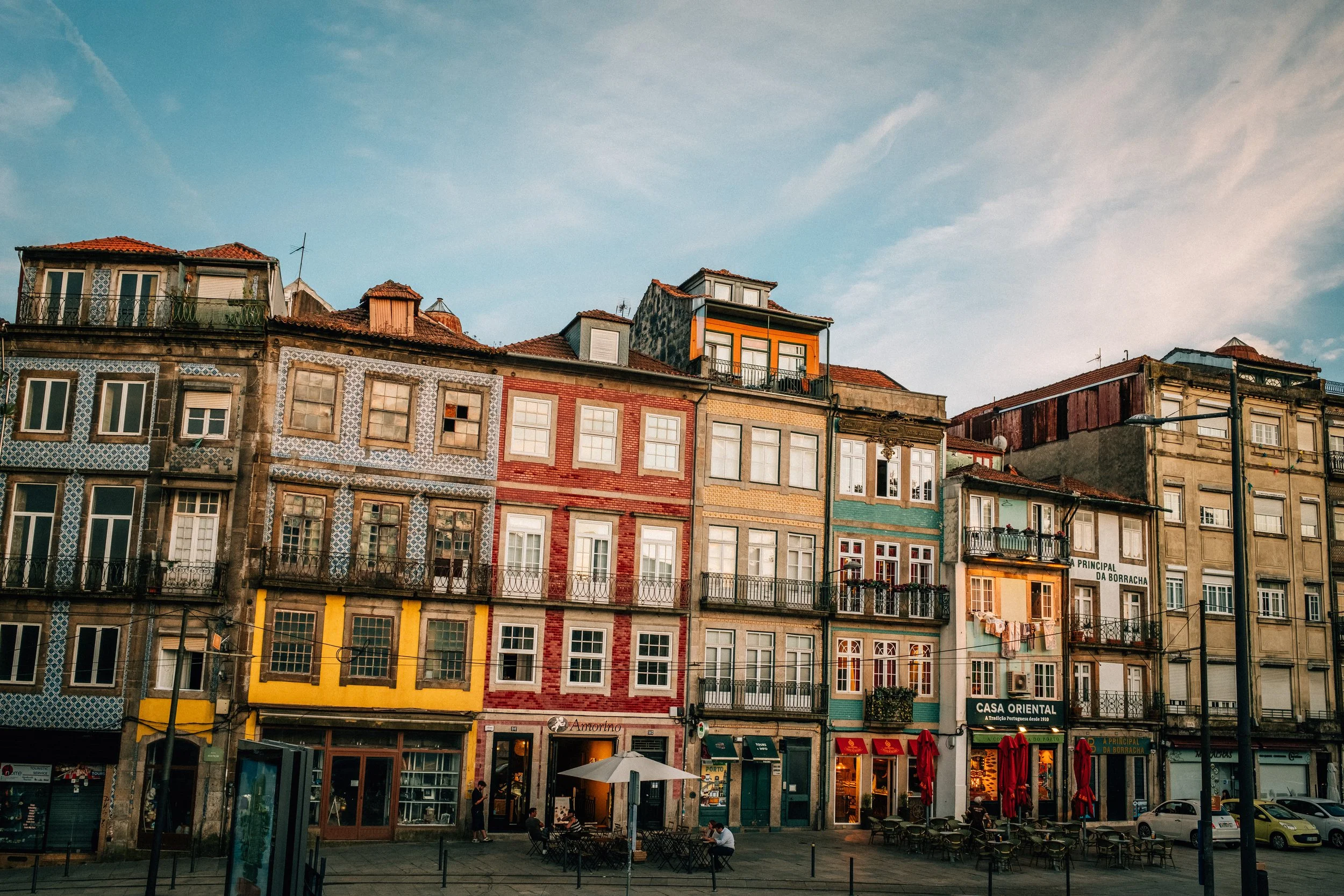 Colorful row of European-style buildings with tiled roofs, balconies, and various window styles, under a partly cloudy sky, with outdoor seating at street level and people walking.