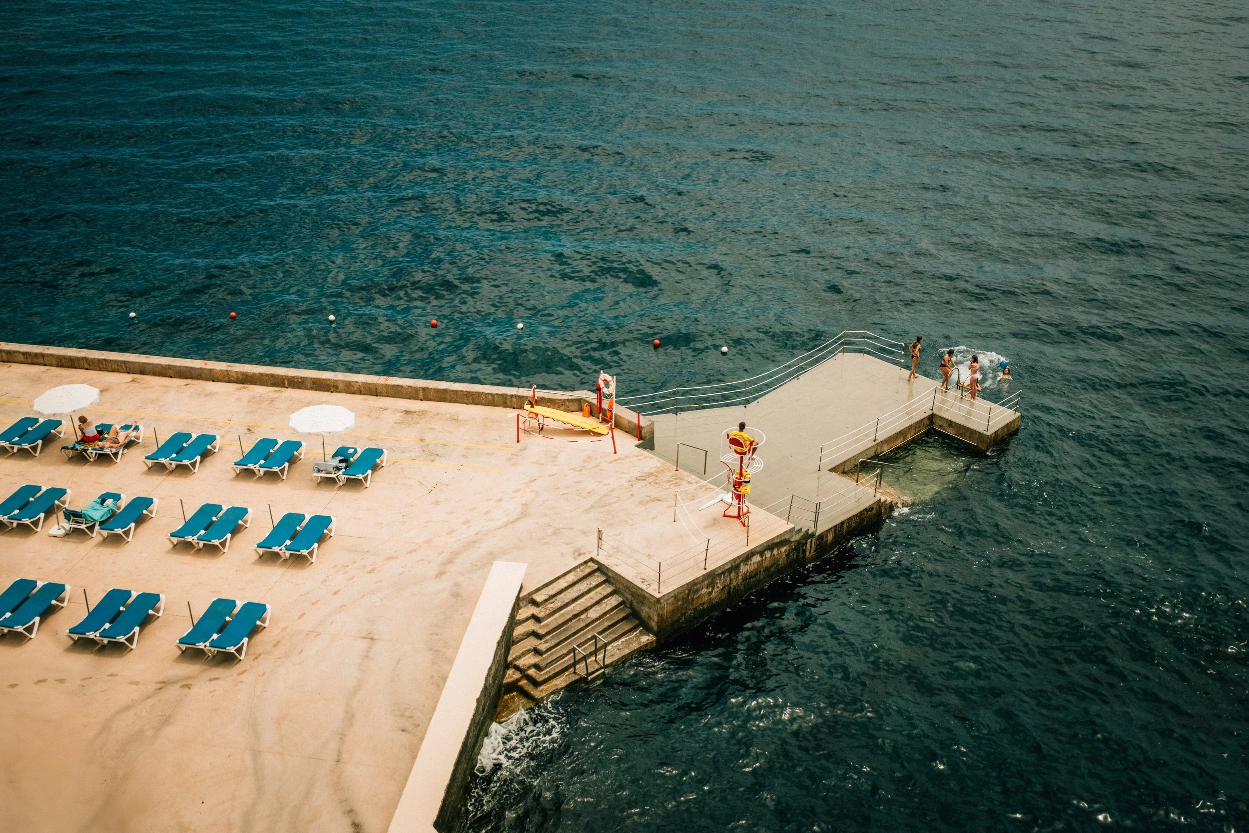 A seaside scene with a concrete dock and stairs leading into the water. Several people are swimming near the dock, and a few are lounging on blue deck chairs under white umbrellas on the sandy beach area adjacent to the dock. The ocean water is deep 