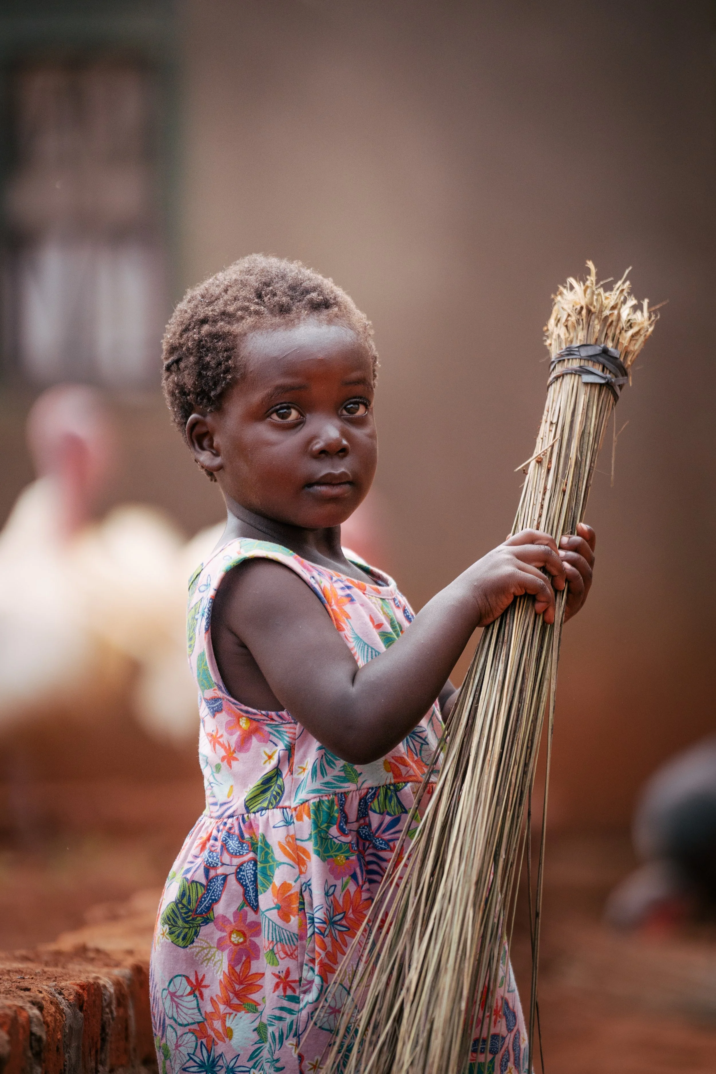 A young girl with short, curly hair holding a bundle of dried grass or reed stalks on a dirt ground, with a blurred background of other children and a brick edge.