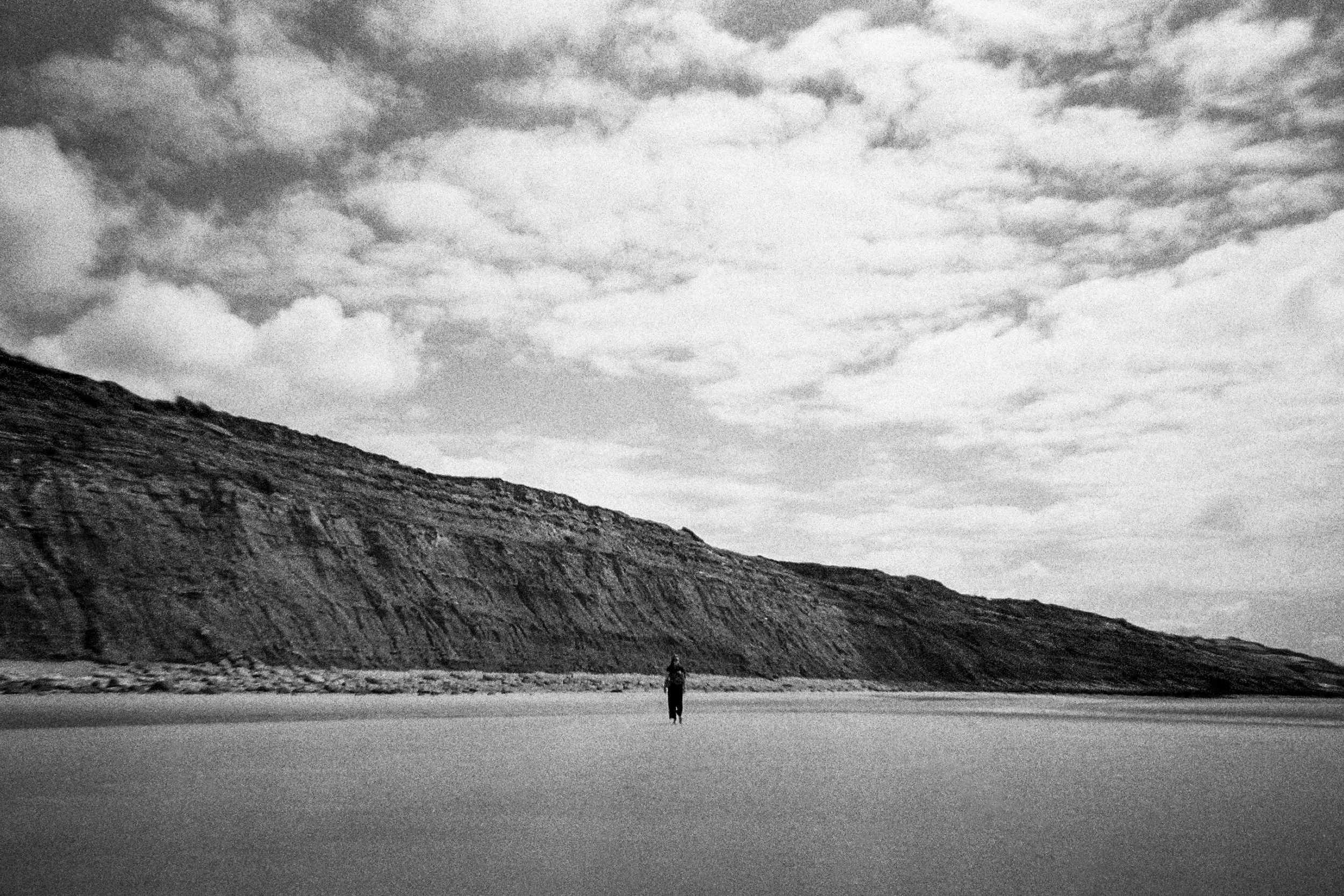 A black and white photo of a person walking on a wide flat landscape with large, steep hills or cliffs in the background and a cloudy sky above.