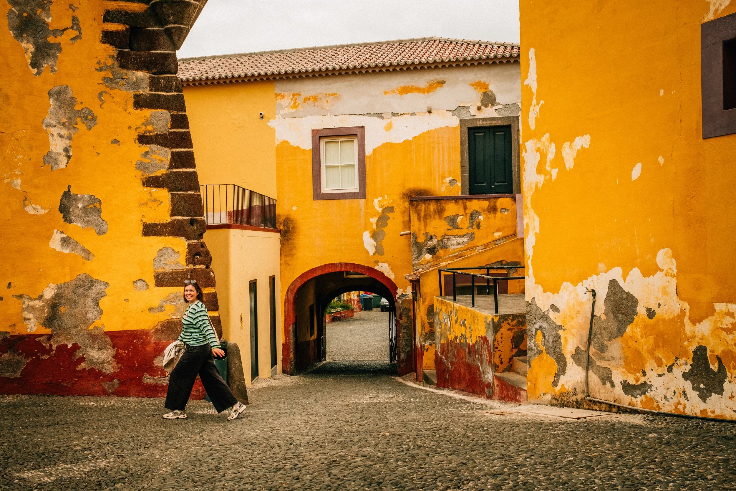 A woman standing on cobblestone street in front of a colorful, weathered yellow and orange building with peeling paint and a dark green door. The building has a second story with a window and a balcony. The sky is overcast.