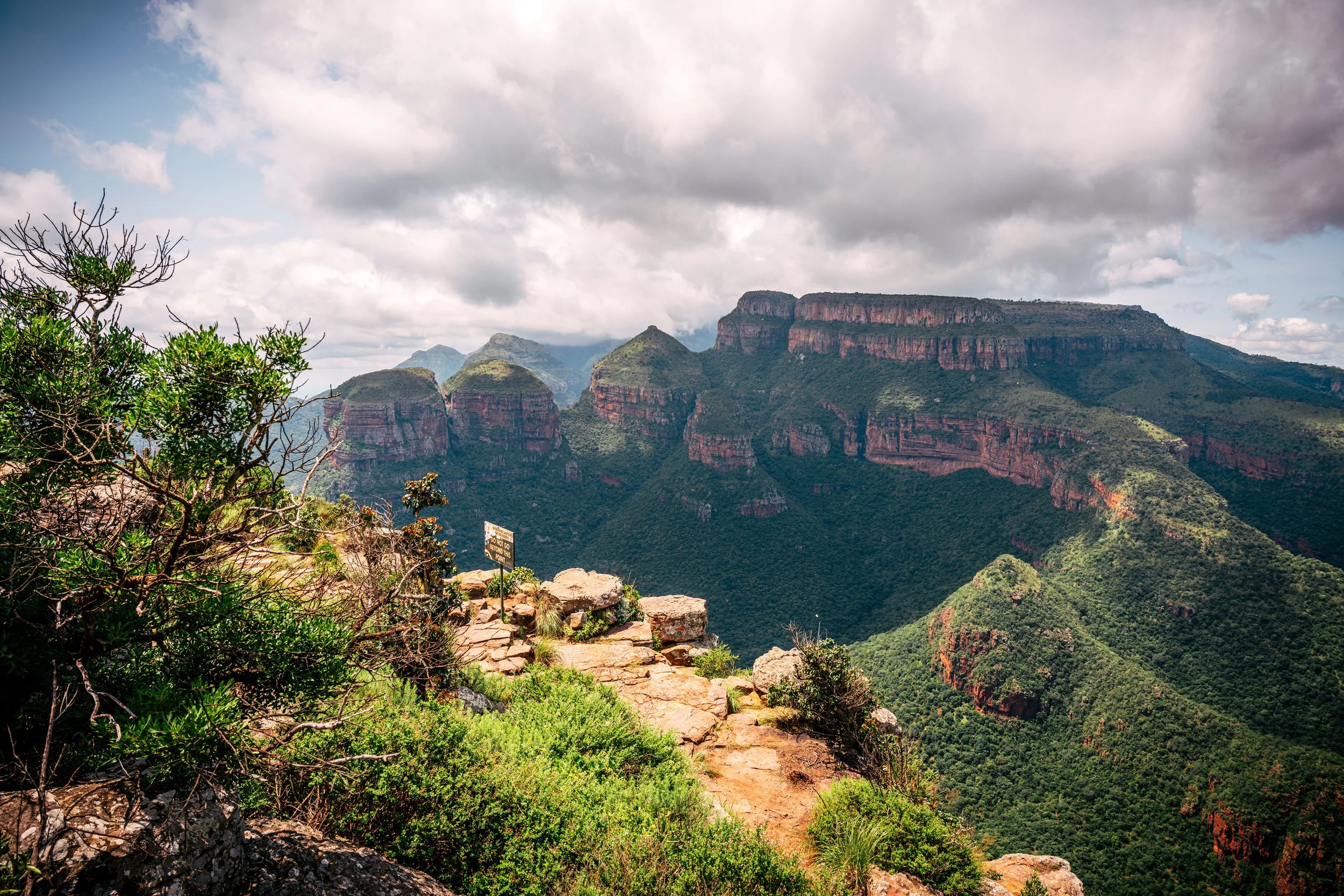 Canyon with reddish rock formations and lush green vegetation under cloudy sky, with a rocky trail in the foreground.