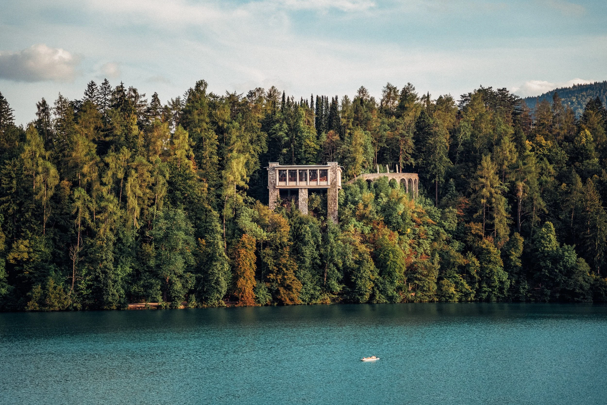 A lake surrounded by a dense forest with a house on a hill and a boat floating on the water.