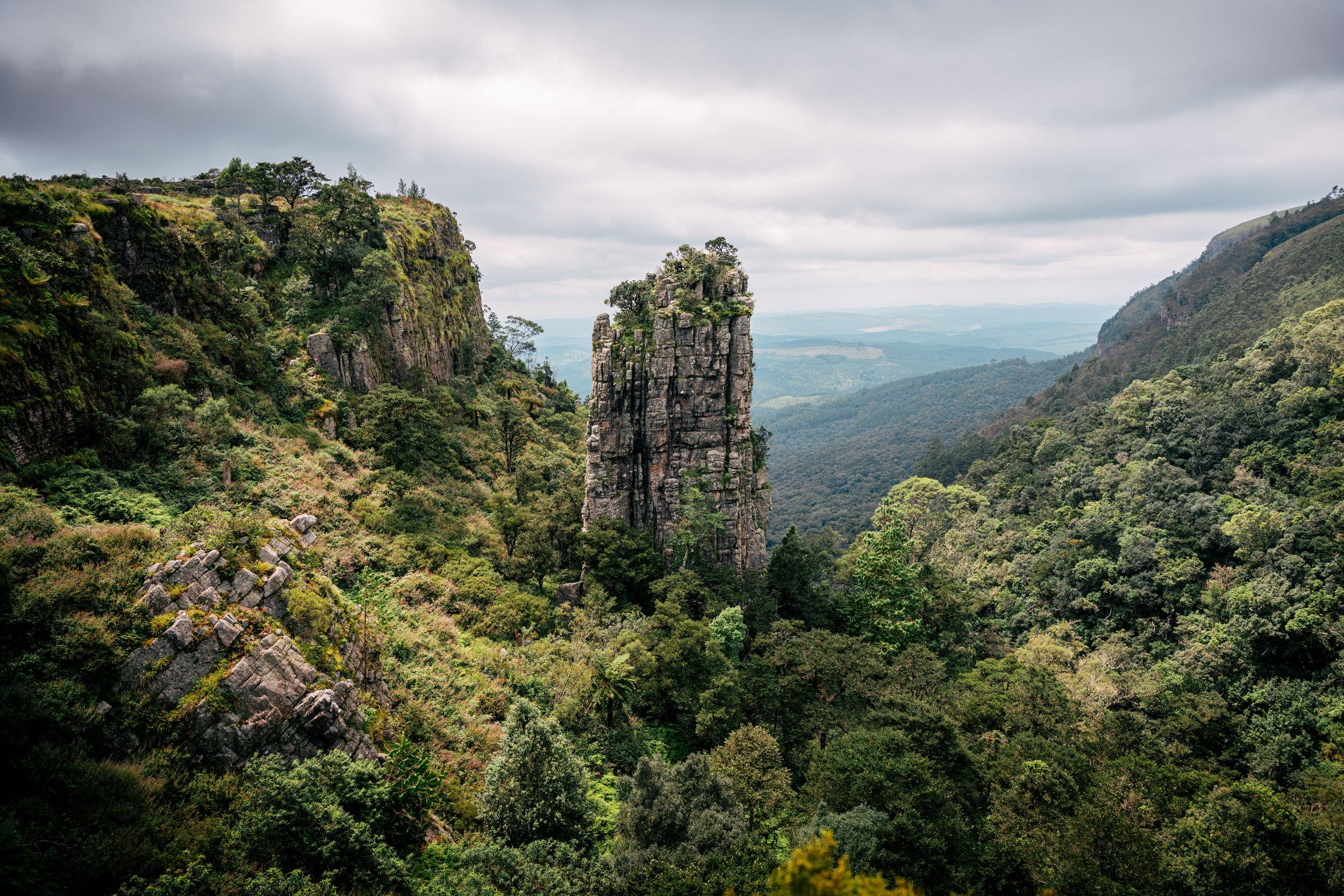 A lush green valley with steep rocky cliffs and a tall, narrow rock formation in the center, under a cloudy sky.