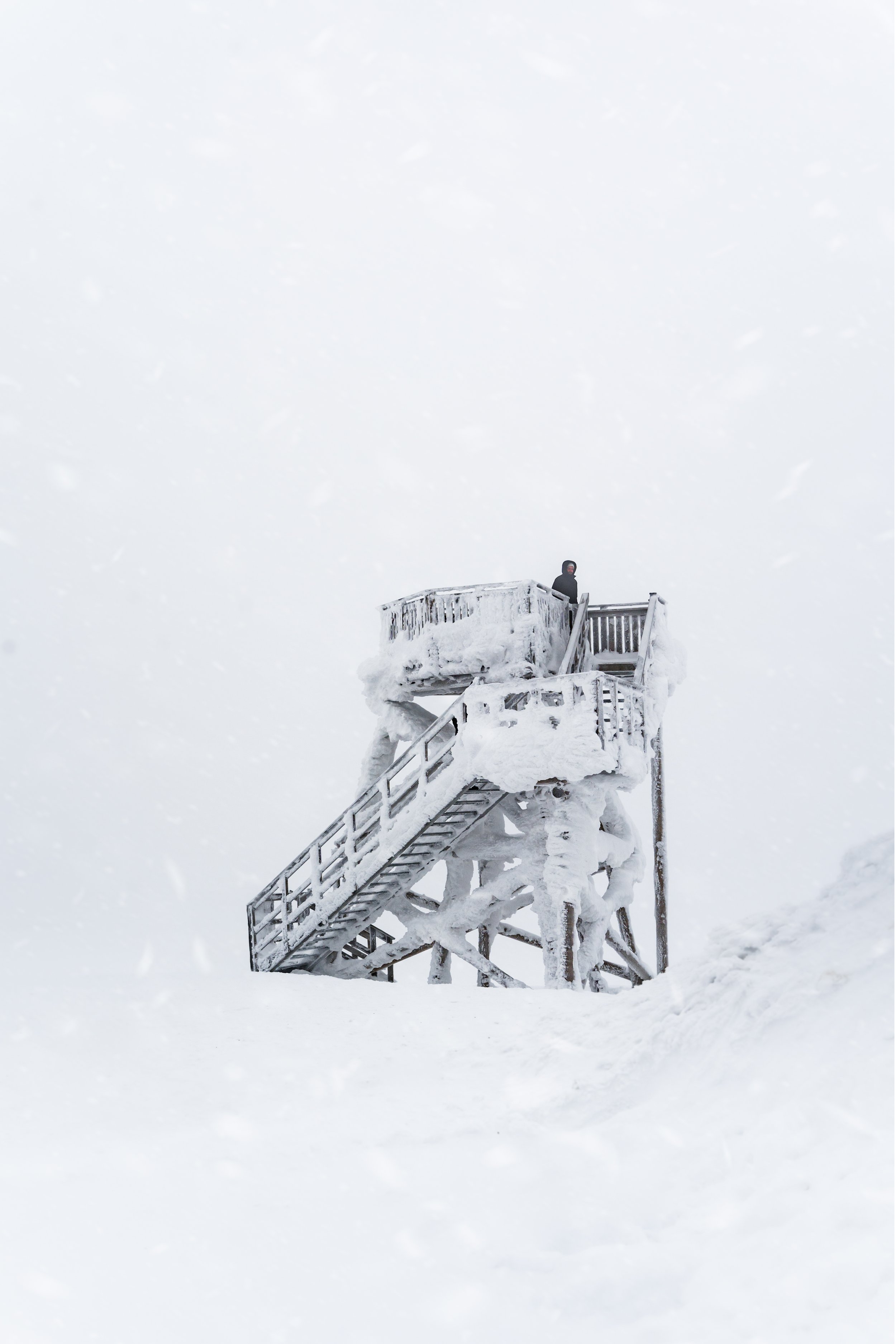 A person in a black jacket and hood standing on a snow-covered observation deck with a staircase, surrounded by a vast snowy landscape.