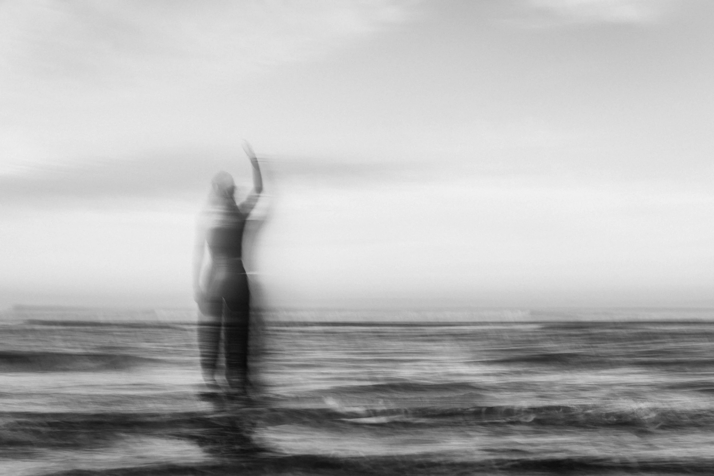 A blurred black-and-white photo of a person standing or dancing on a beach with their arm raised. The background shows a calm sea and sky.