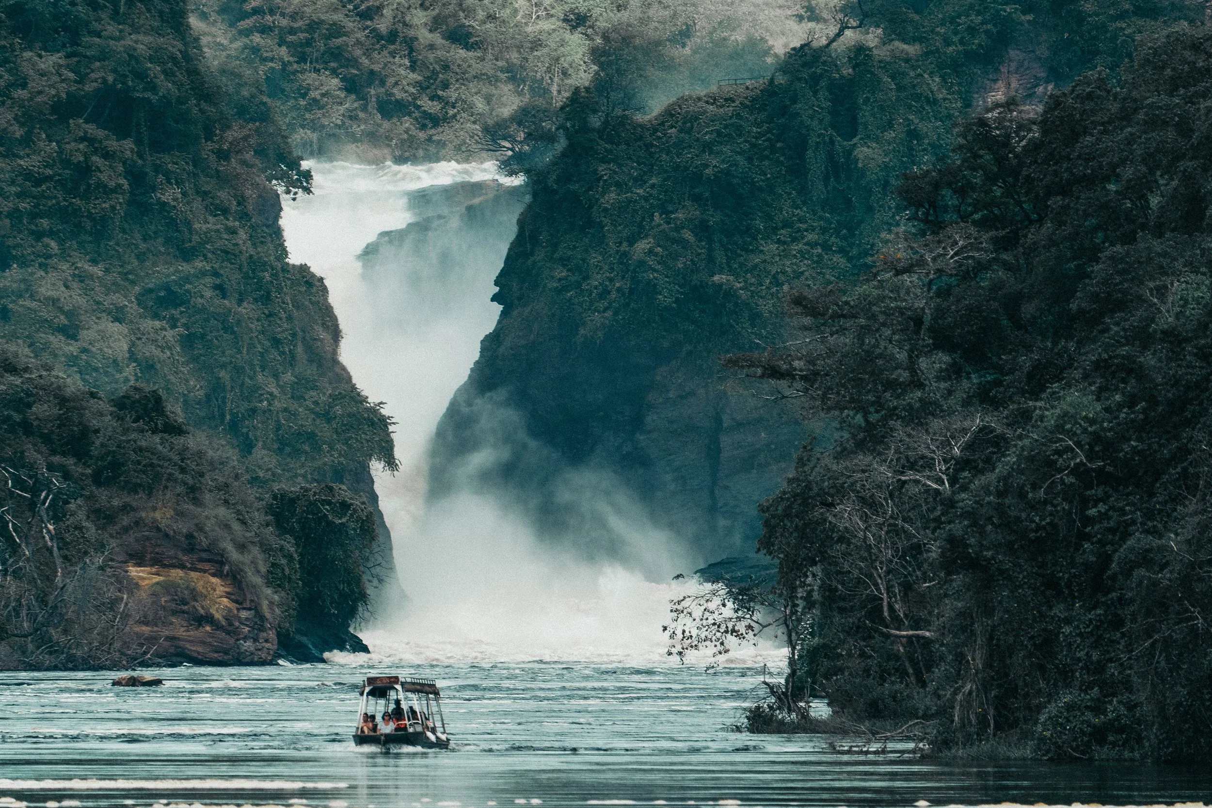 A boat with people on it floating in a river surrounded by lush green trees and a large waterfall in the background.