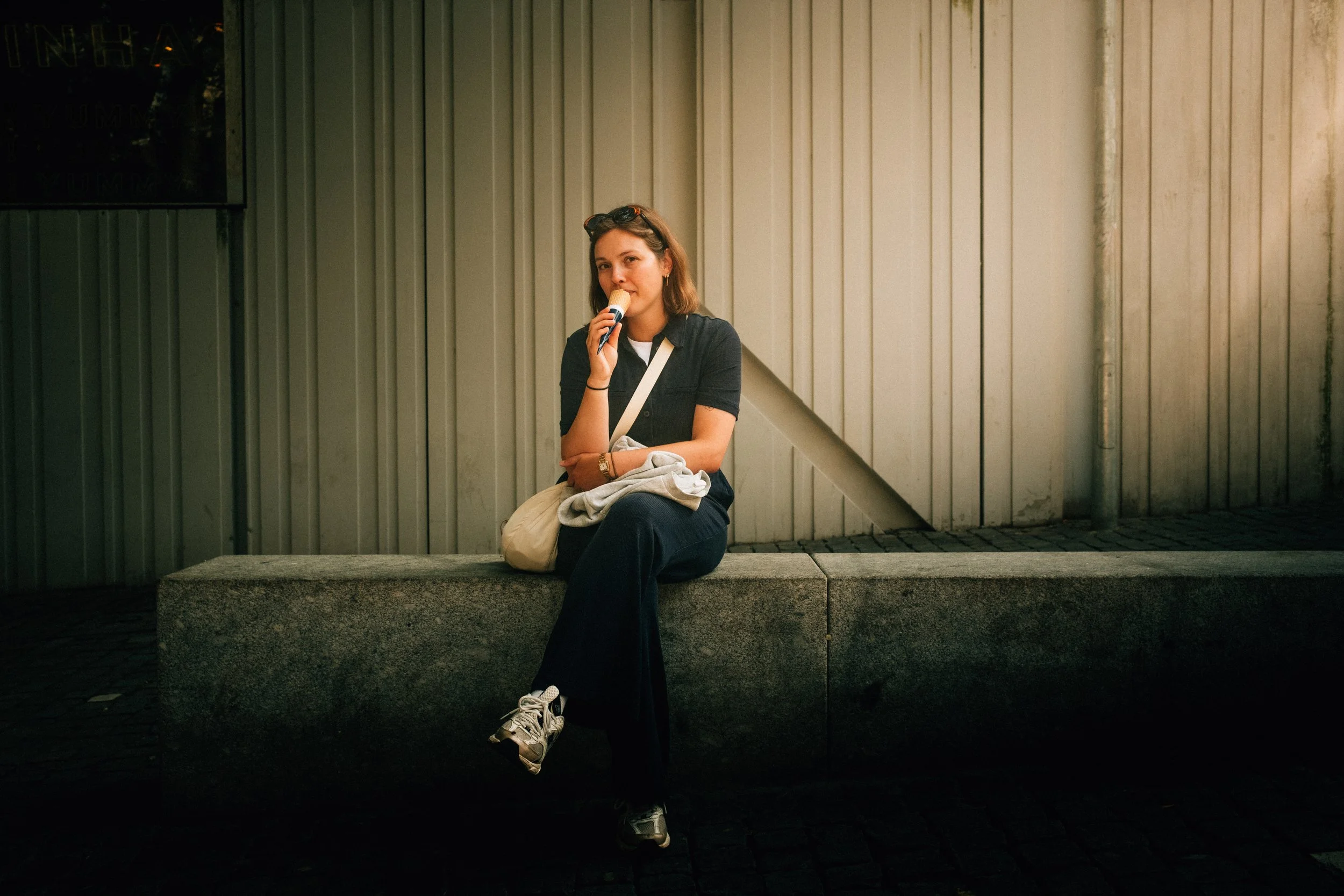 A young woman with shoulder-length brown hair sitting on a concrete bench against a green siding wall, holding an ice cream cone, wearing a black shirt, black pants, and sneakers, with sunglasses on her head and a tote bag on her lap.