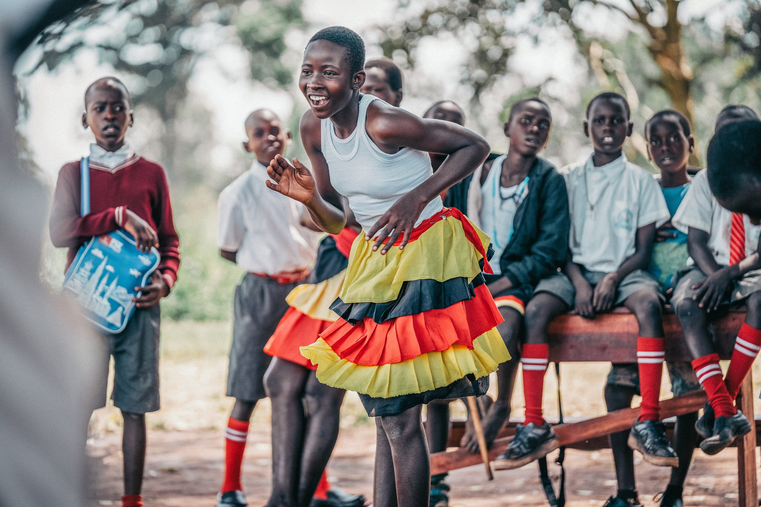Young girl performing a traditional dance in a colorful skirt, surrounded by other children watching outdoors.