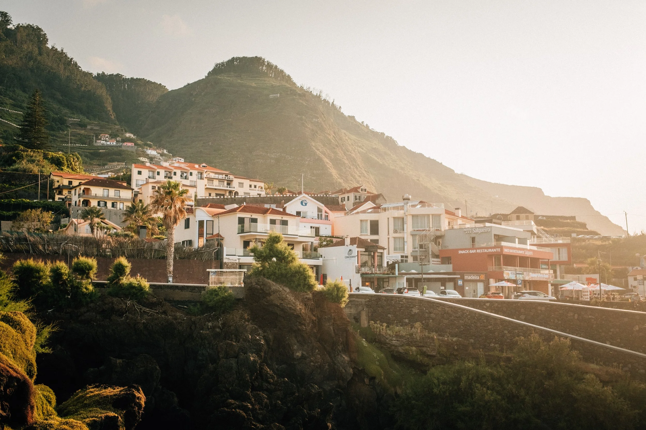 Coastal town with white buildings and red-tiled roofs at the base of a green hillside, with sandy beach and ocean in the background.