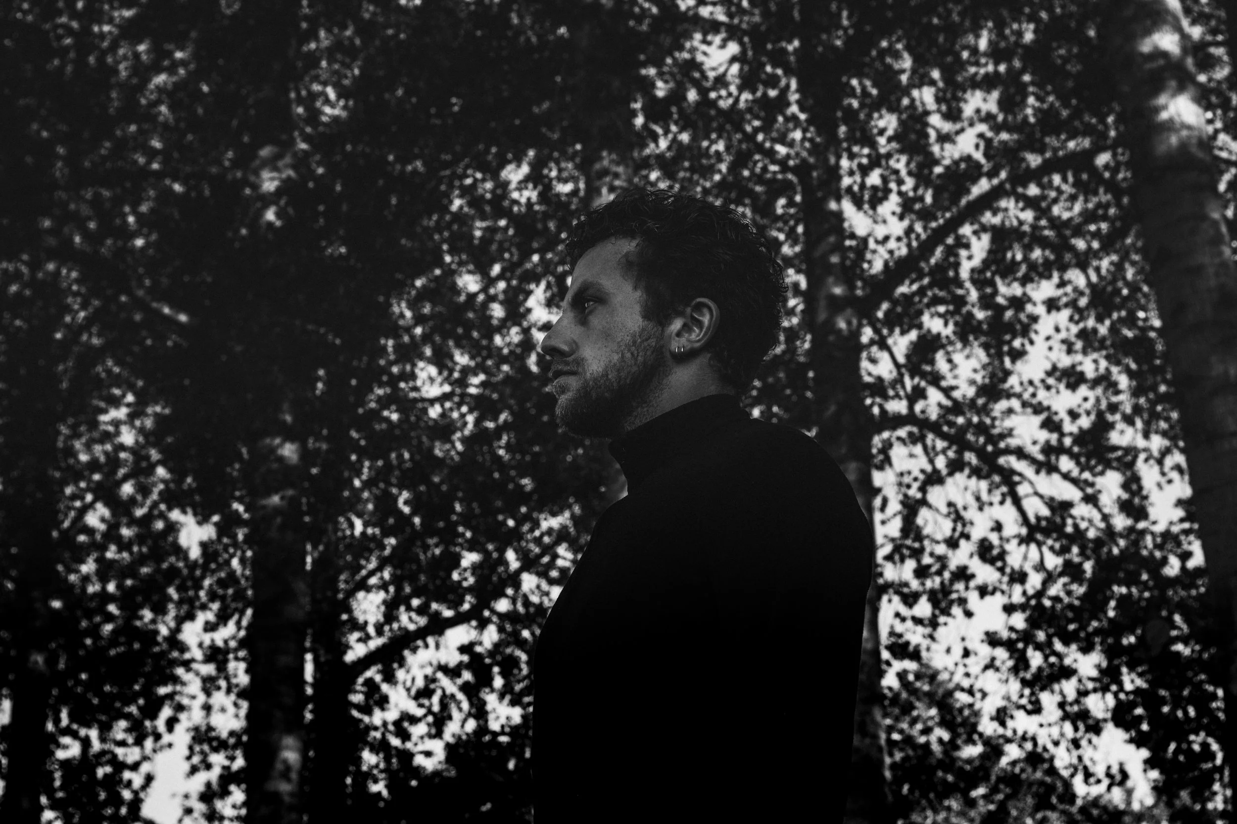 A black and white photo of a man with curly hair and a beard, standing outdoors surrounded by trees, looking to the side.
