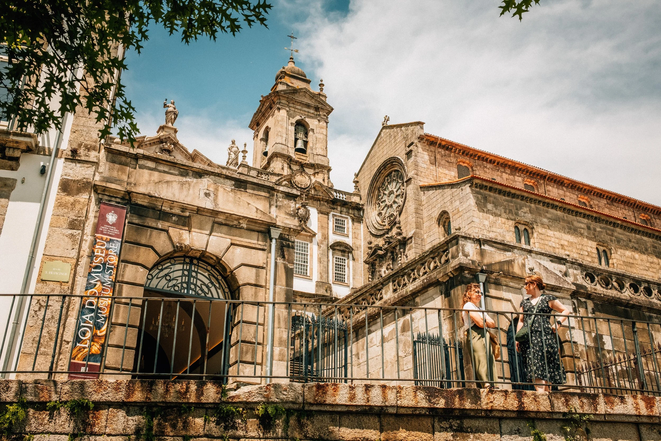 An old historic stone church with ornate architectural details and statues, seen from the street with people talking outside, under a partly cloudy sky.