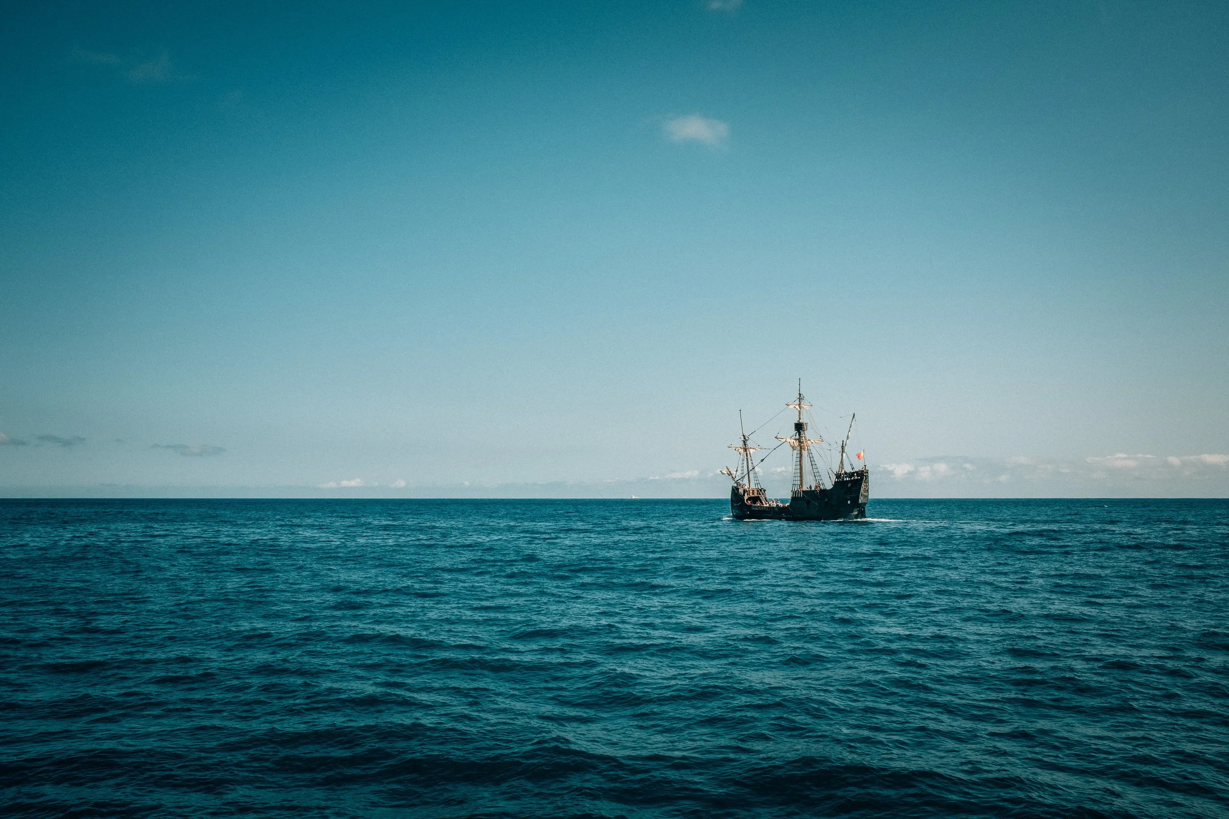 A sailing ship with multiple masts sailing on the ocean under a blue sky with few clouds.