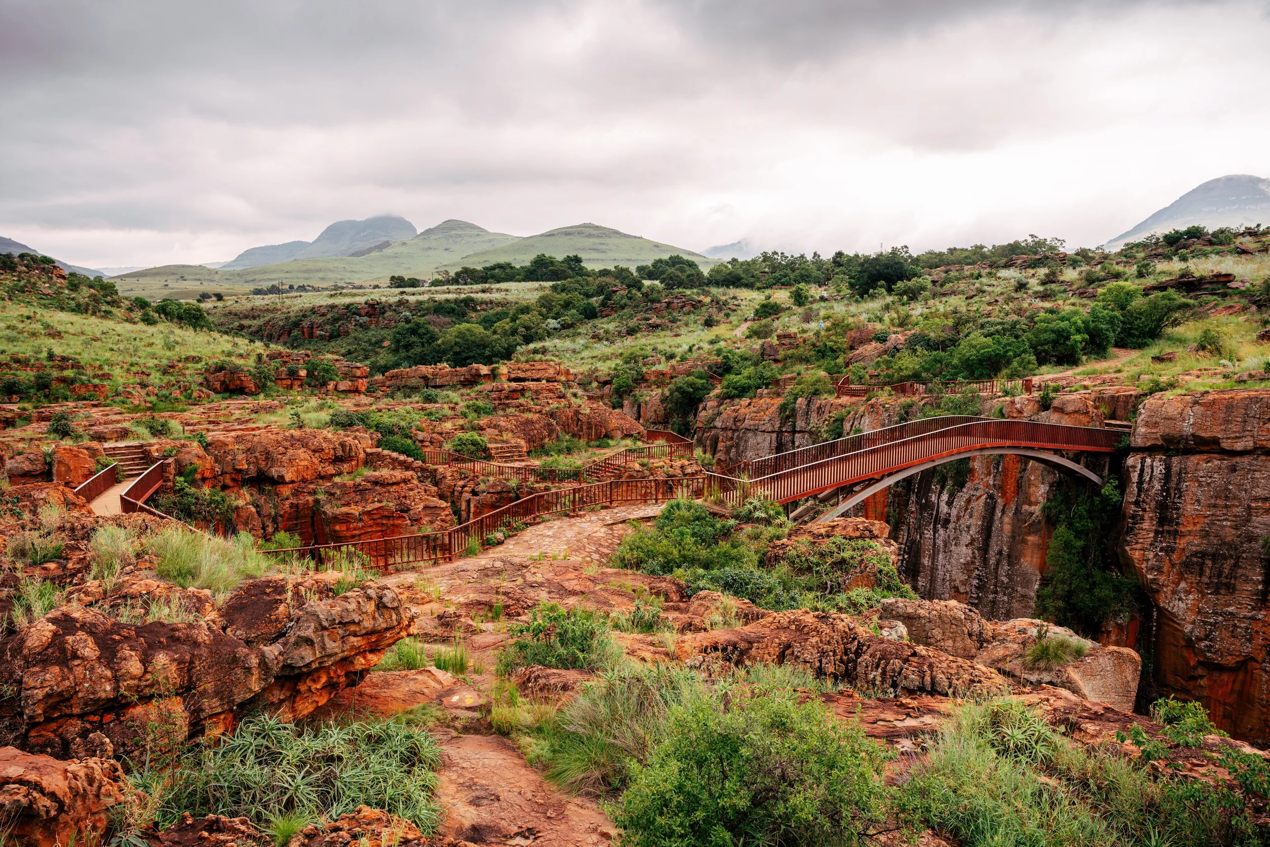 Scenic view of a rocky canyon with red rocks, surrounded by green vegetation, a bridge crossing over the canyon, and rolling hills and mountains in the background under cloudy skies.