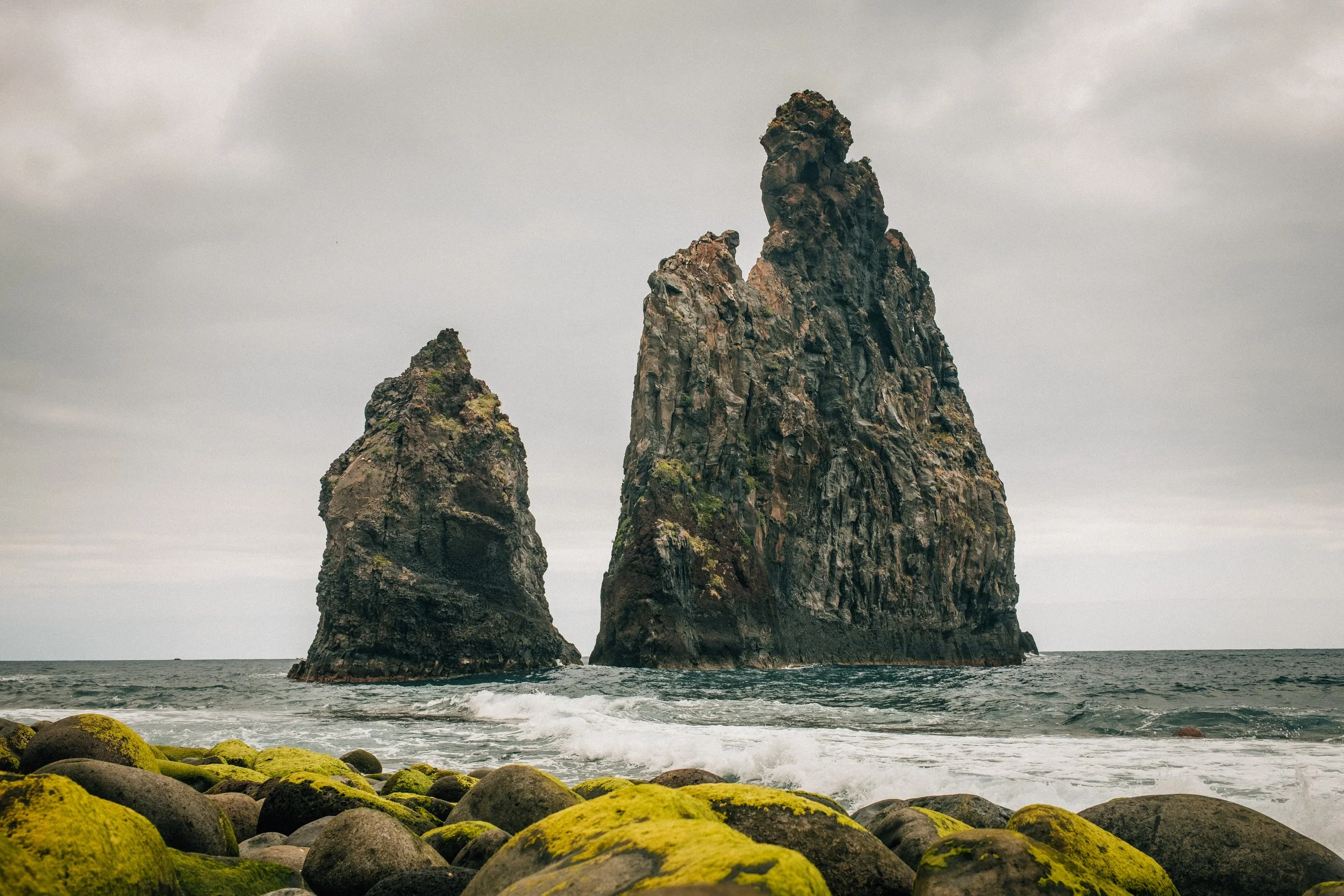 Two large sea stacks rising out of the ocean near a rocky shore covered in mossy stones; overcast sky.