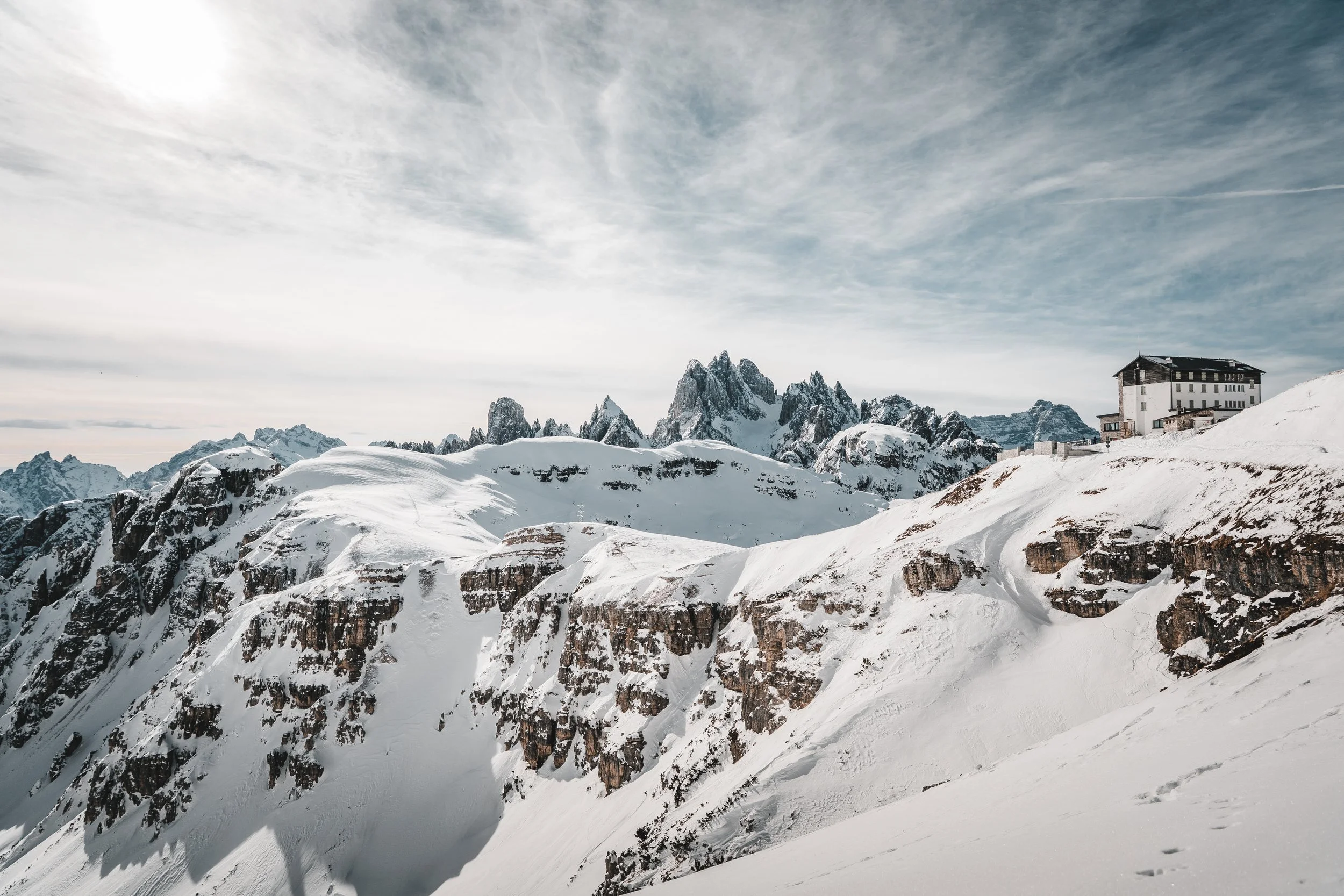 Snow-covered mountain range with jagged peaks and a building on the right side, under a partly cloudy sky.