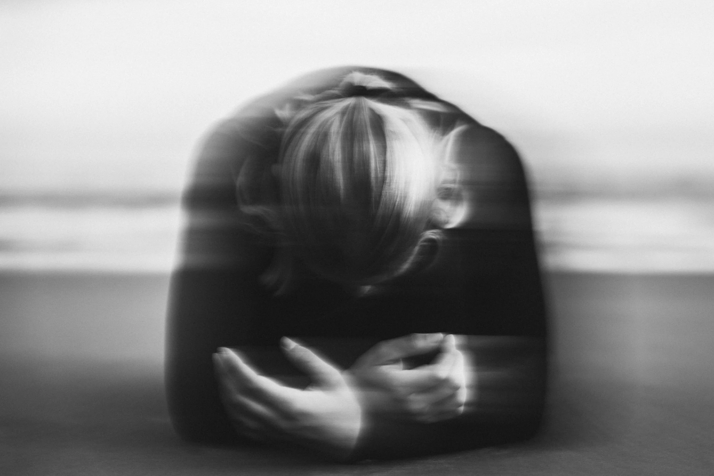 A person with light-colored hair in a bun is leaning over a table, inspecting a clear glass bowl or container. The image is black and white and slightly blurred, with a plain background.