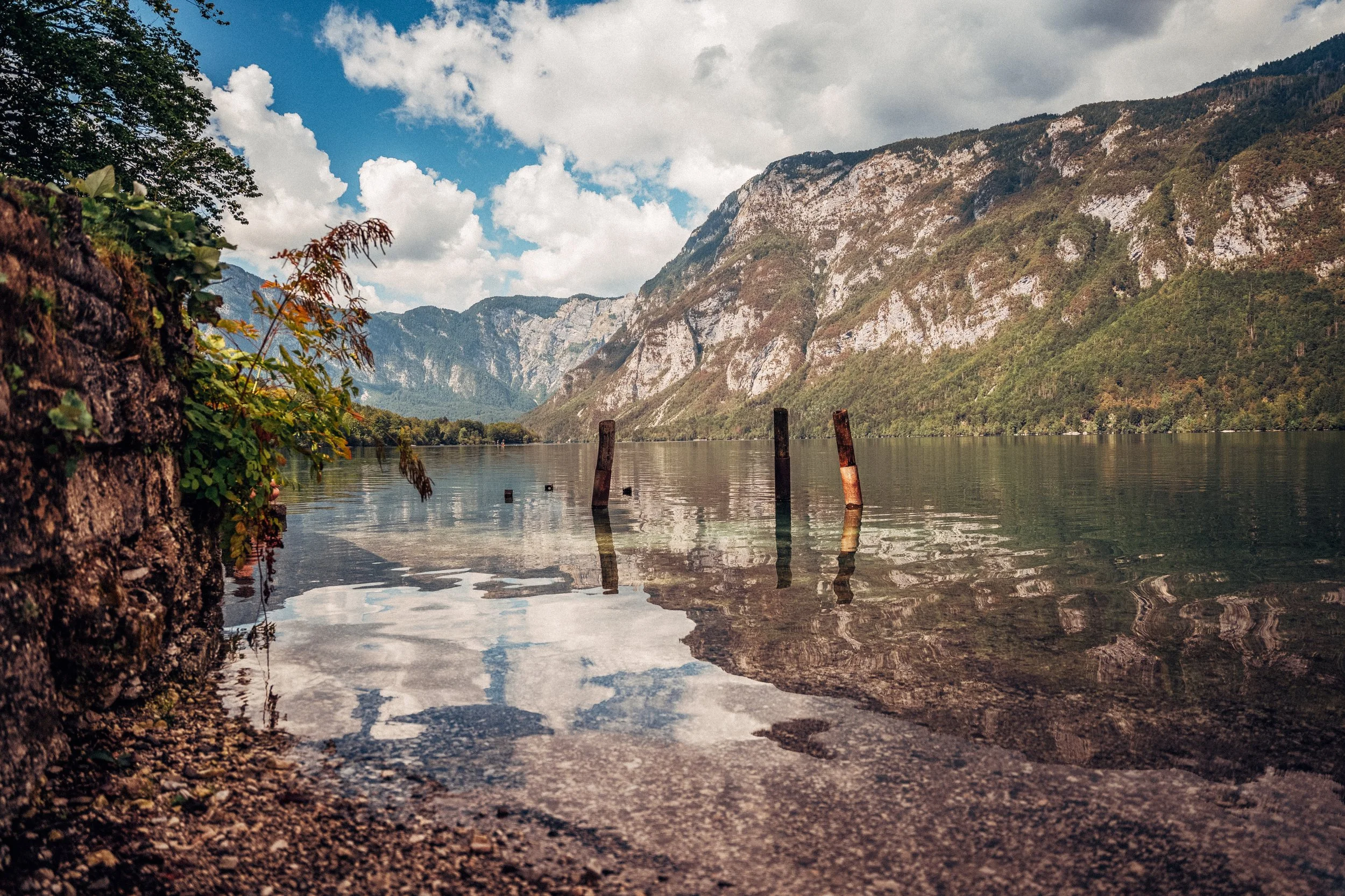 Mountain lake scene with tall mountains in the background, partly cloudy sky, and posts sticking out of the calm water in the foreground.